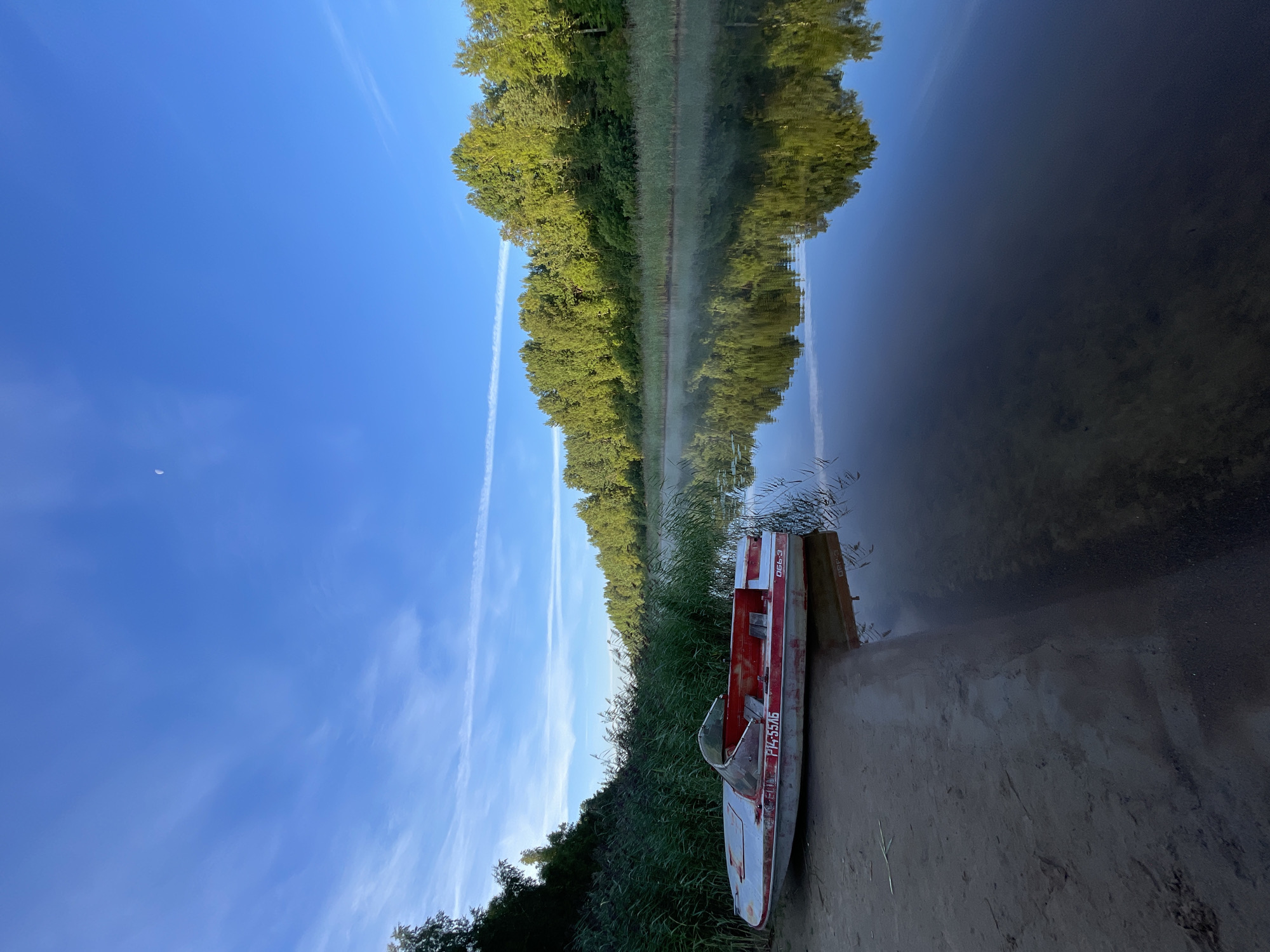 An old, red and white motorboat rests on a sandy shore next to a calm river. The boat is positioned on the left side of the frame, with its bow facing the viewer and its stern extending towards the water. The river stretches across the right side of the image, reflecting the lush green trees that line its banks. A light mist hovers over the water's surface, adding a serene atmosphere. The sky above is a clear blue with a few wispy white clouds and a sliver of a crescent moon visible. The scene is set in Kobona, Russia.