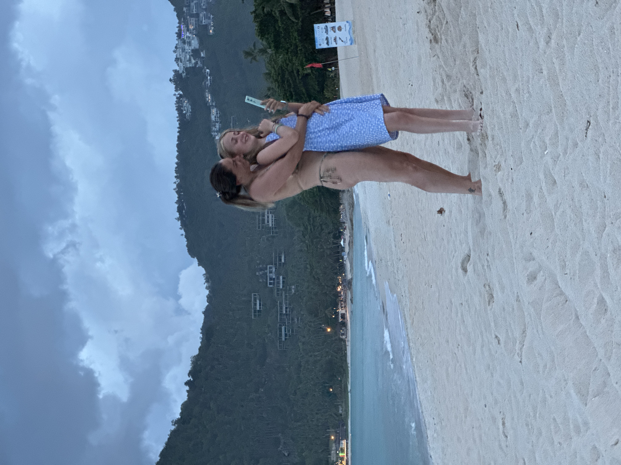 In Baan Chaweng Noi, Thailand, a woman in a bikini embraces a young girl in a blue dress on a sandy beach. The woman is holding the girl from behind, and they are both looking at a smartphone that the girl is holding. The sky is cloudy and the sea is calm with gentle waves. In the background, a lush green hill is visible, dotted with white buildings. There is also a signpost and some beach chairs on the sand. The overall scene suggests a peaceful and intimate moment between two people enjoying their time by the sea.
