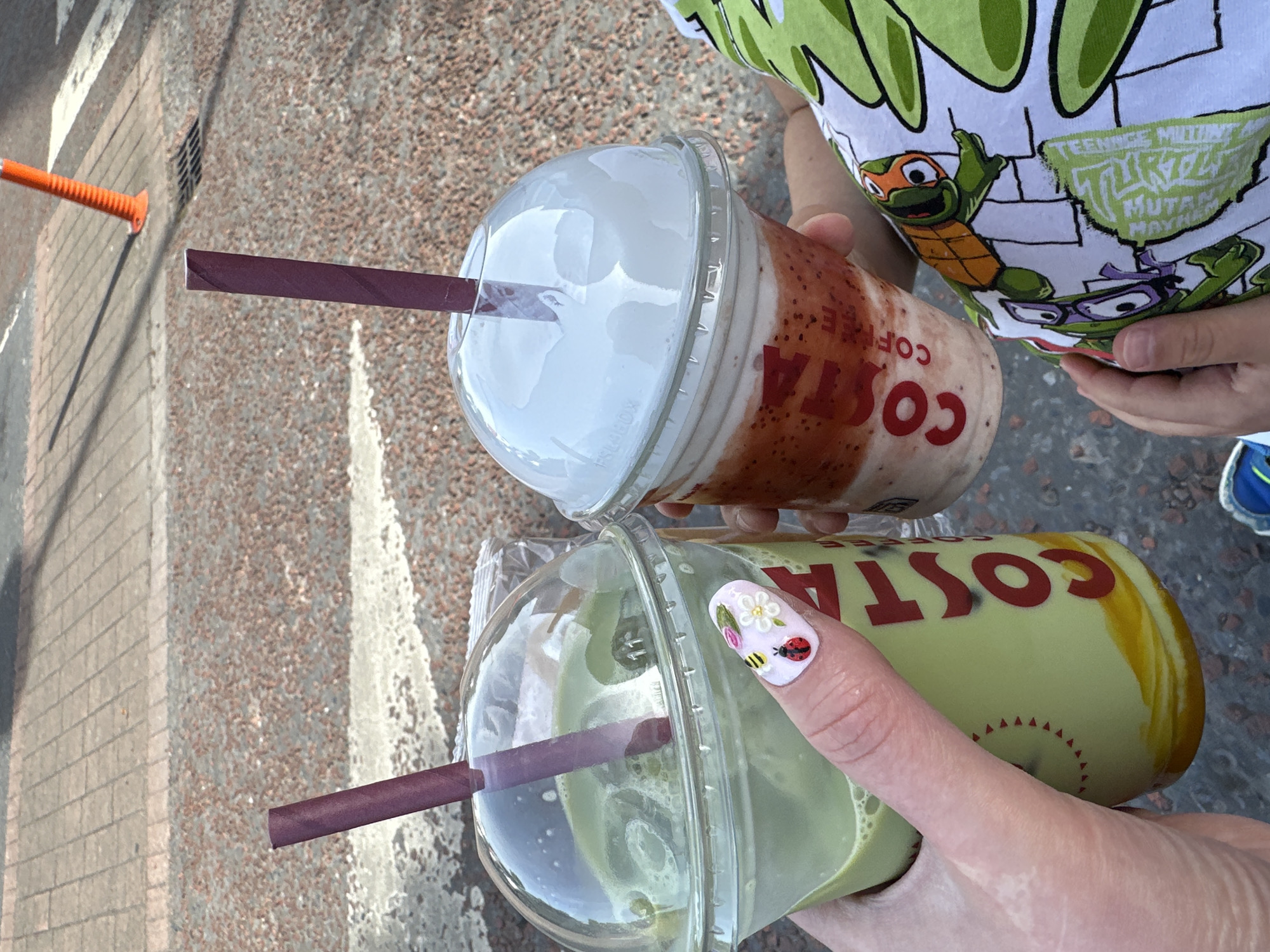 In Carlow town, Ireland, two Costa Coffee drinks are held by individuals. The drink on the left is a green matcha beverage in a clear plastic cup with a dome lid and a purple straw. A hand with decorated fingernails, featuring a ladybug, bee, and flowers, is holding this cup. The cup has red "COSTA" lettering on its side. To the right, a strawberry-flavored drink in a similar cup with a dome lid and a purple straw is visible. This drink has visible layers of strawberry and cream. The background shows a textured ground of small red pebbles and grey paving stones, with a bright orange traffic cone in the upper right corner. A person wearing a green shirt with a Teenage Mutant Ninja Turtle print is partially visible on the right side of the image, with their hand reaching towards the strawberry drink.