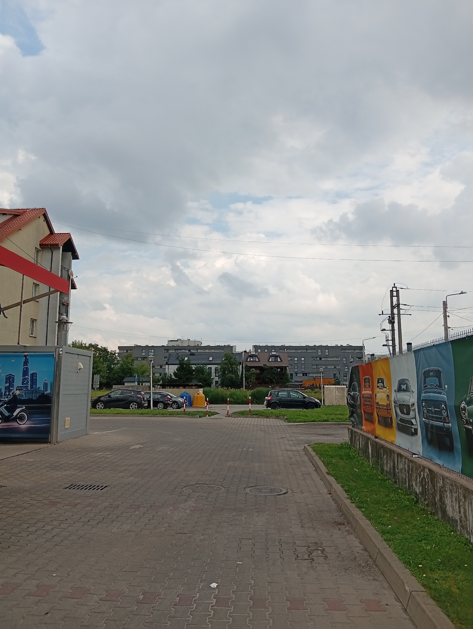 The image shows a street scene in Błonie, Poland, on a cloudy day. In the foreground, a paved walkway runs alongside a grassy area bordered by a concrete wall. The wall is decorated with a colorful banner featuring images of vintage cars. To the left of the walkway, there is a building with a red roof and a beige facade, and a gray structure with a large advertisement on its side. Several cars are parked on the street in the middle ground, and in the background, there are modern apartment buildings and trees. Power lines and utility poles are visible against the sky.