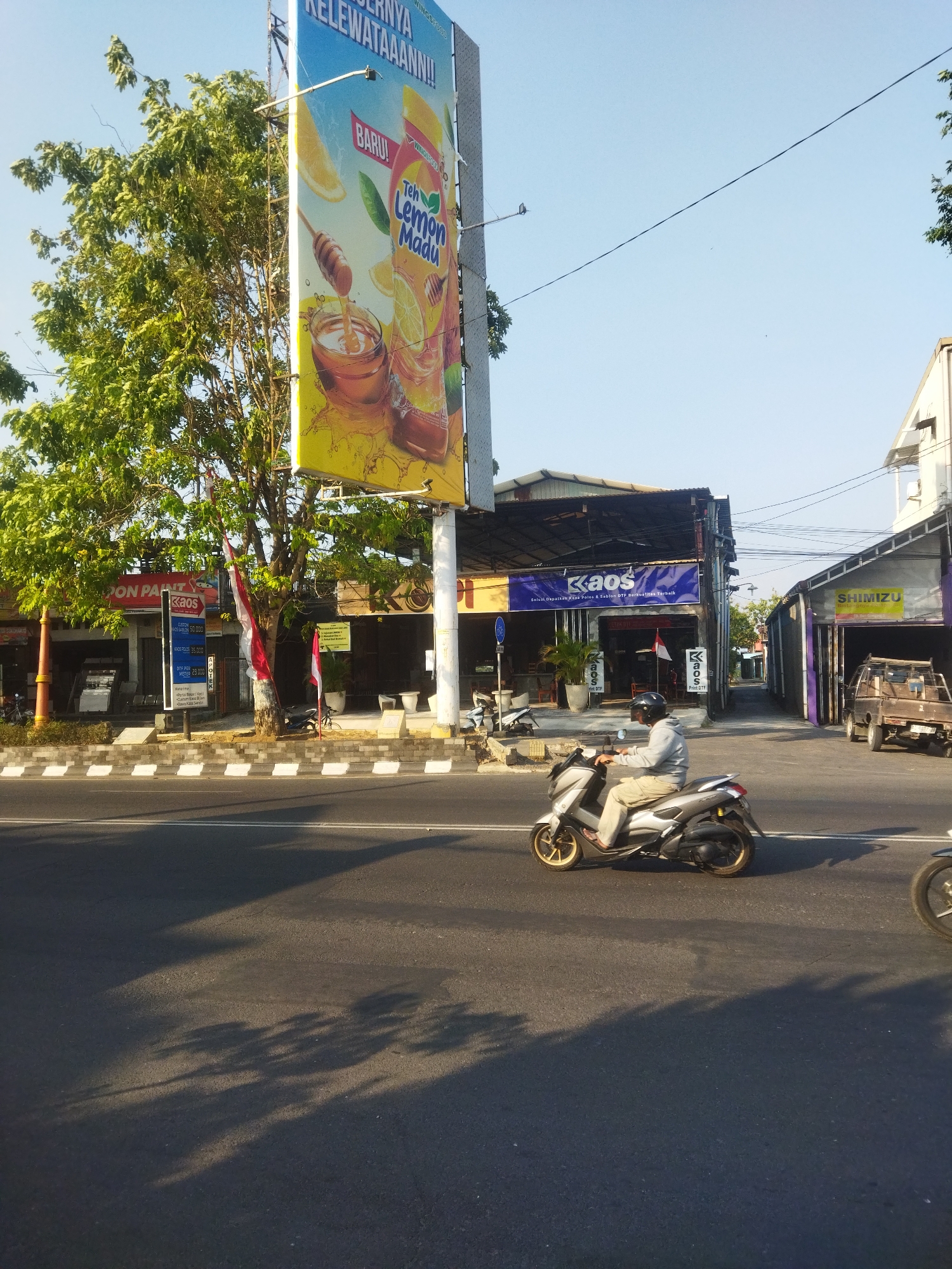 The image depicts a street scene in Sukoharjo, Indonesia, with a focus on a large billboard advertising "Teh Lemon Madu" (Lemon Honey Tea). The billboard is situated on the right side of the road and features a colorful illustration of lemons, honey, and a beverage. In the foreground, a person is riding a silver scooter with gold rims along the asphalt road. The scooter rider is wearing a gray long-sleeved shirt, light-colored pants, and a helmet. To the left of the billboard, a large tree with green foliage is prominent, partially obscuring some storefronts. Several shops are visible in the background, including one with a blue and yellow banner that reads "KAOS" and another with a yellow sign featuring the word "SHIMIZU." Indonesian flags are displayed in front of some of the buildings. The sky is clear and blue, indicating a daytime scene. The road is marked with white lines, and shadows of trees are cast on the asphalt, suggesting the sun is positioned to the left of the frame.