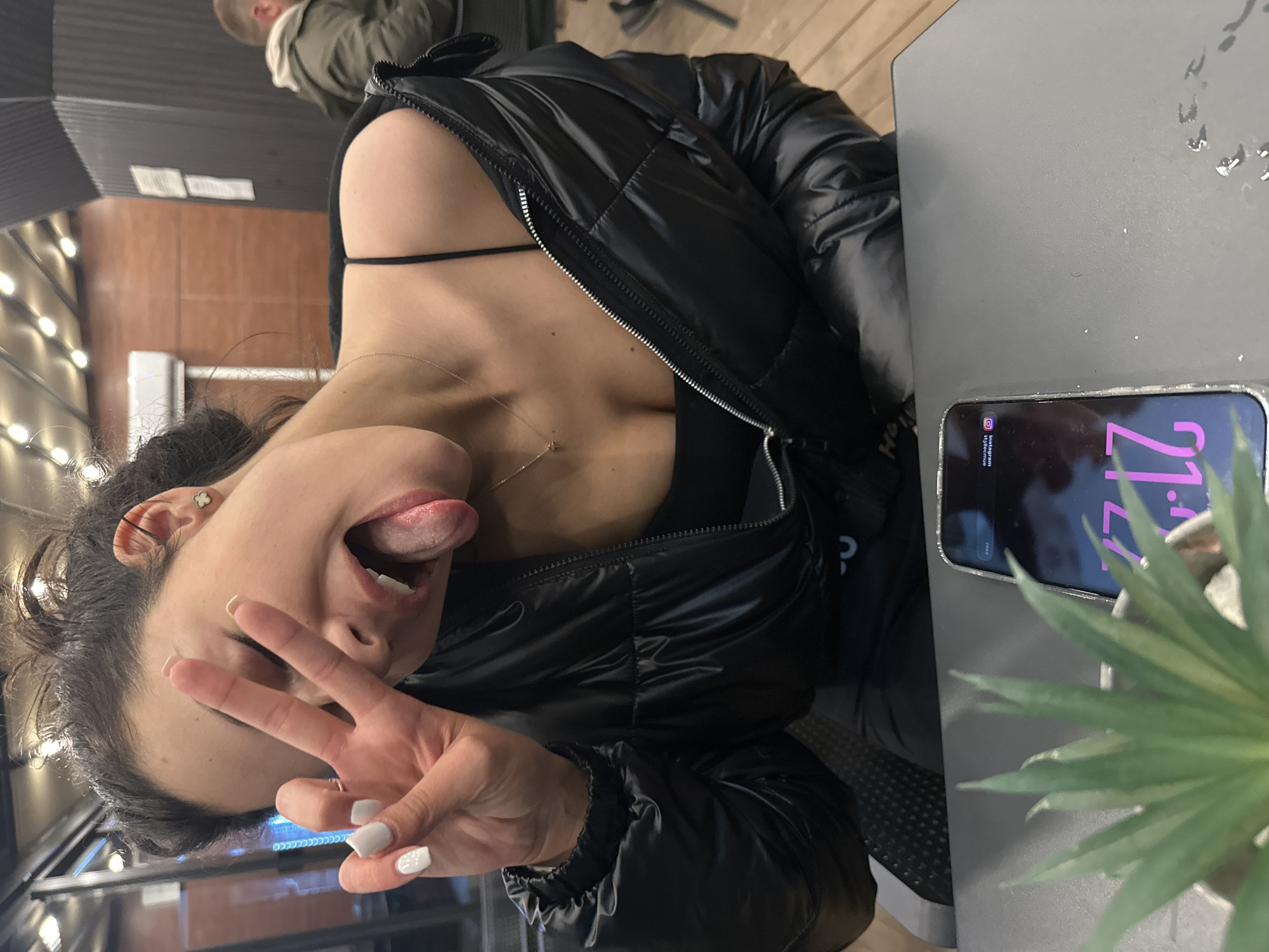A young woman poses at a table outdoors, sticking out her tongue and making a peace sign with her hand. She wears a black jacket and a low-cut top, with a gold necklace visible. A smartphone rests on the table next to a small potted plant, displaying "21:17" on the screen. The location is Plovdiv, Bulgaria, as indicated by the "Instagram Bulgaria" label on the phone screen. The setting appears to be a cafe or restaurant patio. Another person is partially visible in the background.