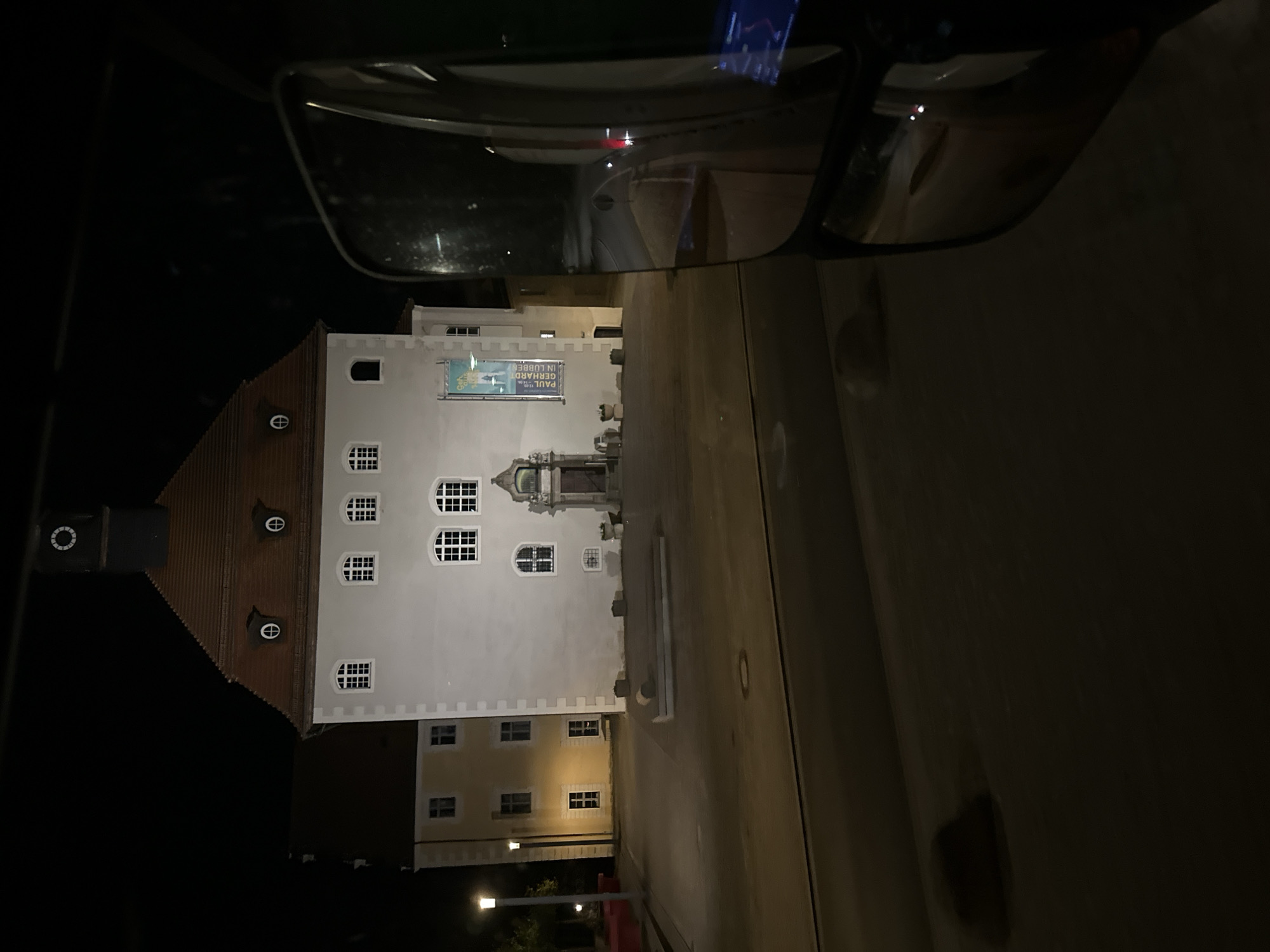 The image shows a building in Lübben (Spreewald), Germany at night, seen from the perspective of a vehicle. The building is a light-colored, multi-story structure with a red tiled roof. It has several arched windows with white grids, and a clock tower on the left side. A banner with text is hanging on the facade. In front of the building, there is a paved area with some benches and planters. The foreground of the image is dominated by a car's side mirror, which reflects a street and more of the building. Streetlights illuminate the scene.
