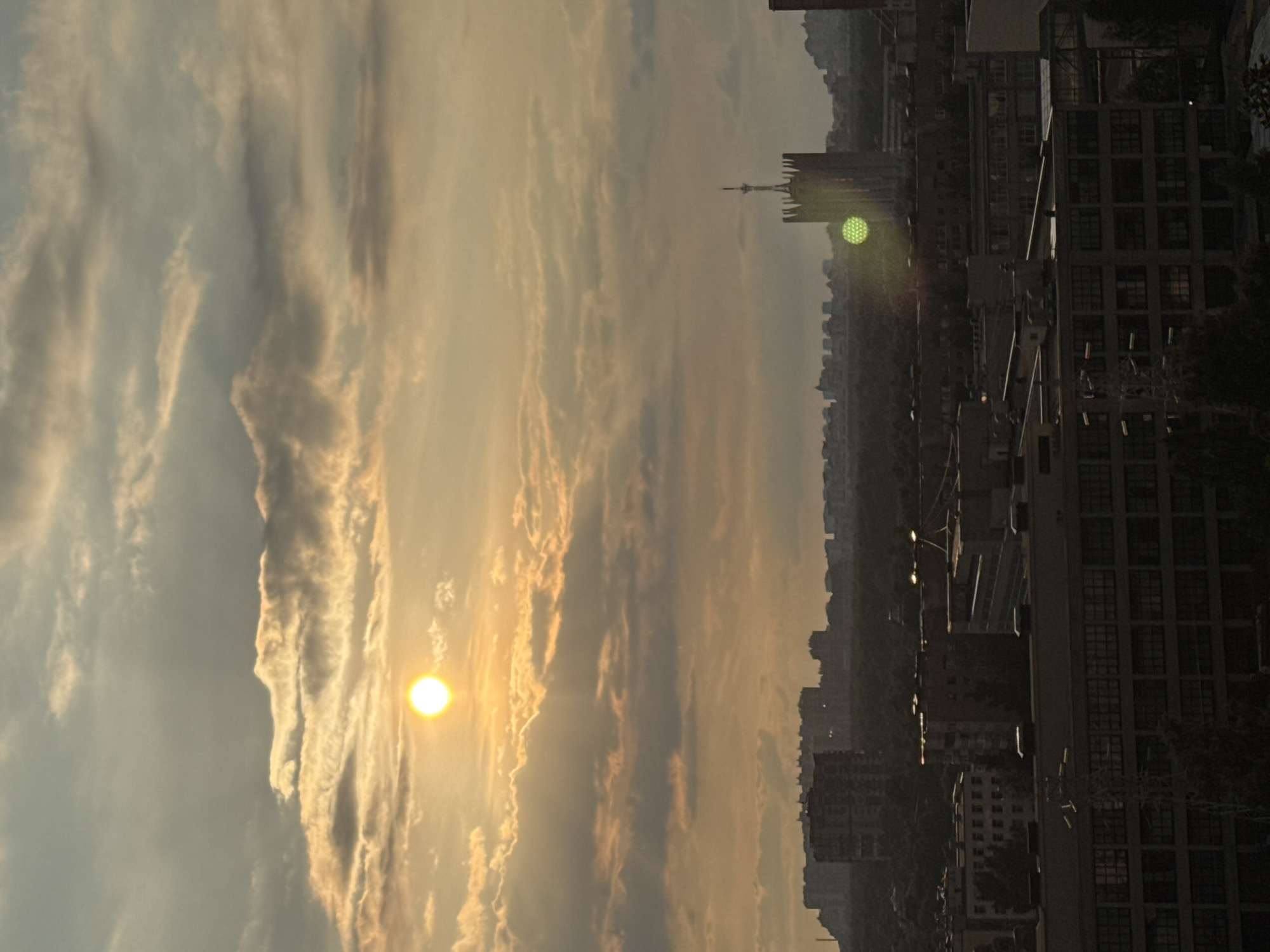 This is a vertical photograph capturing a sunset over a city, likely Sinyavino, Russia. The sun, a bright, small, yellow orb, is partially obscured by wispy, elongated clouds that are illuminated by its golden-orange light. The sky transitions from a pale blue at the top to a warm, hazy orange and yellow near the horizon.
To the right of the sky, a city skyline is visible. Numerous buildings of varying heights, mostly dark and silhouetted against the sky, are densely packed. Some buildings appear to be residential or office blocks with many windows, while others are taller structures, including what looks like a communication tower. A noticeable green circular light or lens flare is present in the middle distance, possibly from a street lamp or another light source. The overall atmosphere suggests an urban evening scene. The time of day is clearly sunset, indicated by the low sun and warm sky colors. The weather appears to be clear, with the clouds being atmospheric rather than indicative of impending rain. No people are discernible in the image. There is no visible text.