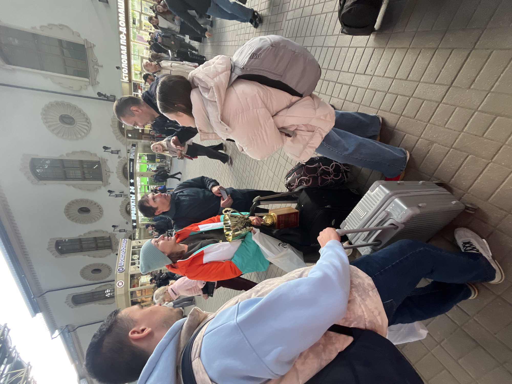 The image captures a bustling scene at the Kazansky railway station in Moscow, Russia. Several individuals are present, many with luggage, suggesting they are travelers. People are engaged in various activities, such as walking, standing, and pulling suitcases. There is a trophy being carried. The station’s architecture is visible, with ornate details on the facade and signage in Russian. The overall atmosphere indicates a busy transportation hub.