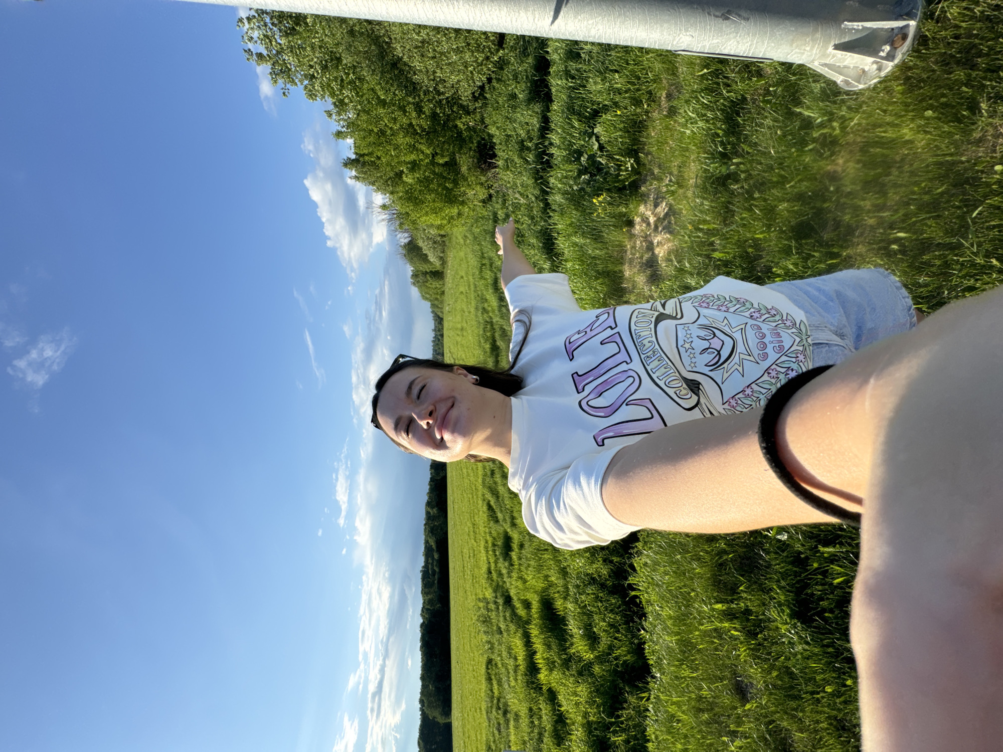 A photograph of a young woman in Bolshiye Vyazyomy, Russia, taken in an outdoor, grassy field area on a sunny day. The image is oriented so the woman is at an angle to the horizon. The woman has her left arm extended out to the left of the frame. She is wearing a white t-shirt with "COLLECTION" written on it and jeans. A cylindrical post is in the upper left of the frame. The sky is blue with sparse clouds. Green trees are in the background.