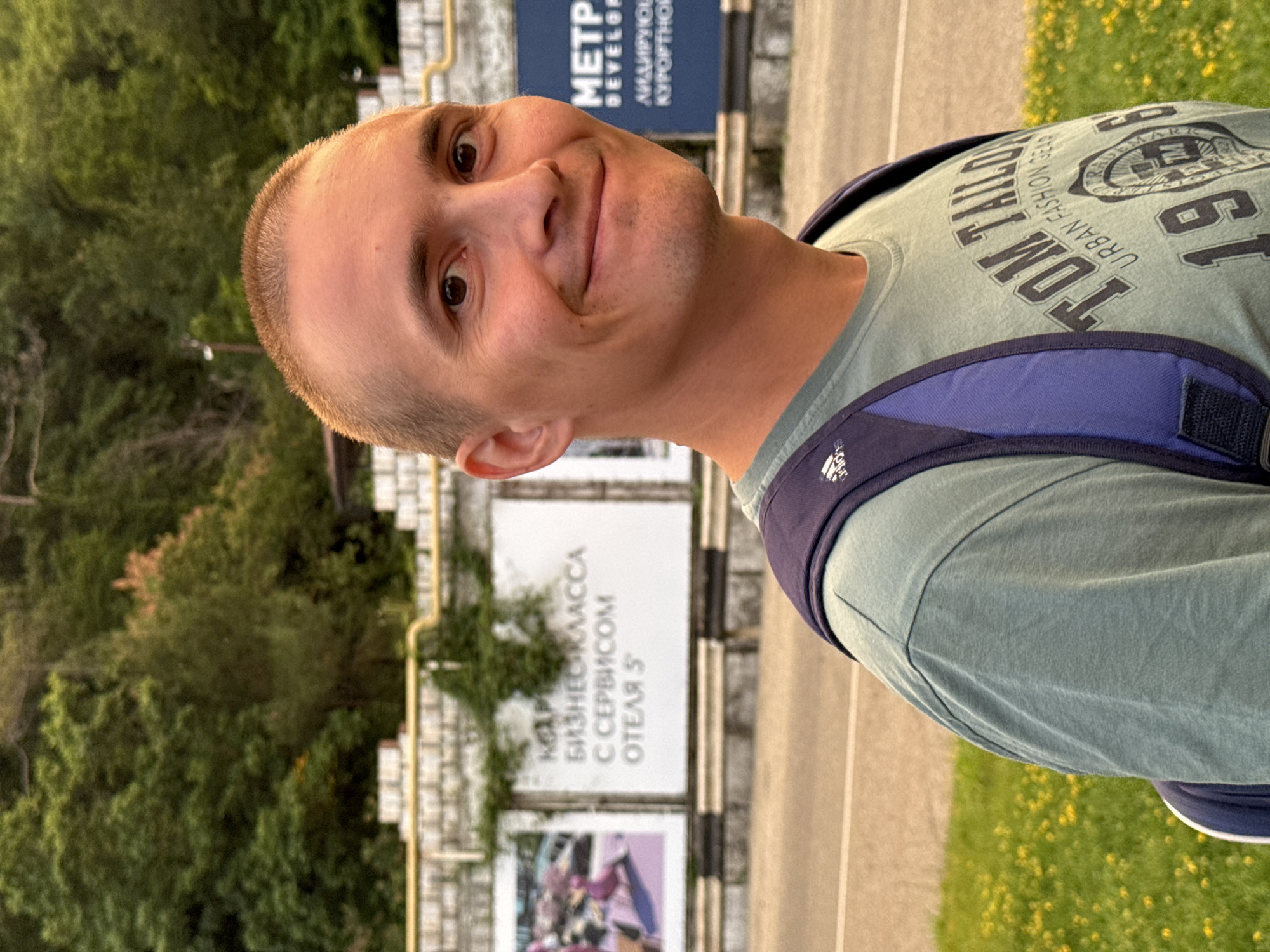A young Caucasian man with a buzz cut and light brown eyes is captured in a medium close-up portrait. He is wearing a light green t-shirt with "TOM TAILOR URBAN FASHION GEAR" printed on it and a dark blue backpack. He is smiling and looking slightly to his right. The background is blurred, showing a street with a yellow line and a grassy area with yellow wildflowers. Behind the street, there are billboards with Russian text and images, and a wall with plants growing on it. Further back, lush green trees form a hillside. This scene is likely located in Nebug, Russia, given the presence of Cyrillic text on the billboards, possibly indicating a resort town on the Black Sea coast.