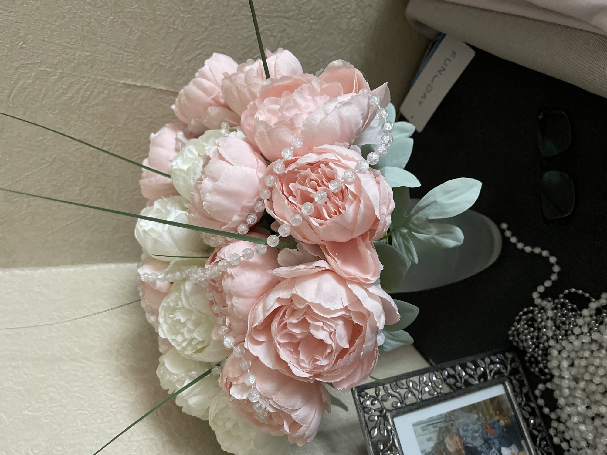 A still-life image shows a bouquet of pink and white artificial peonies adorned with beaded strands. The bouquet is arranged in a light-colored vase, which is placed on a dark surface. Near the vase are eyeglasses and a partial view of a picture frame, along with a cascade of beads. The image does not provide any information about the city of Moscow or people.