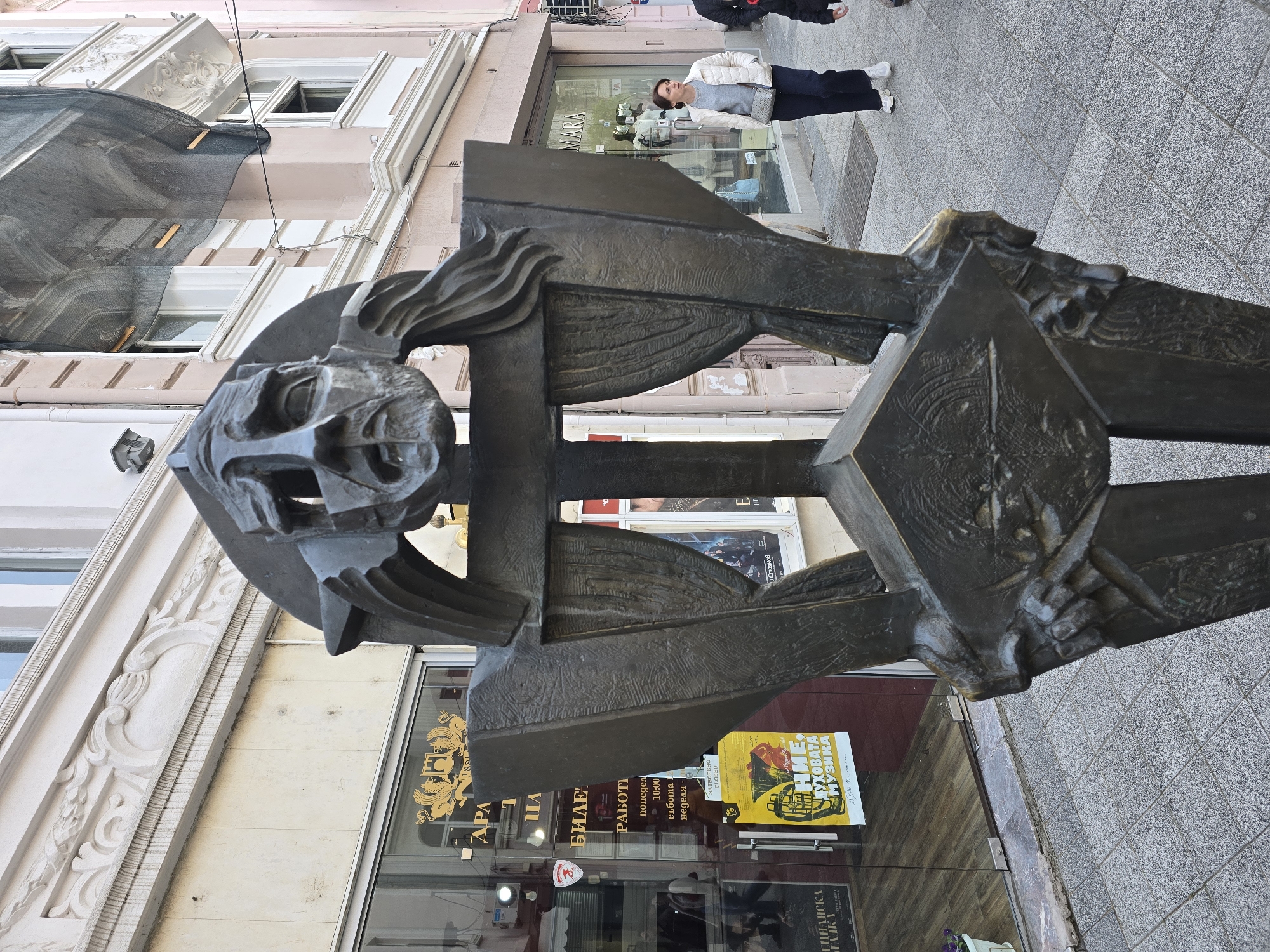 A ground-level, outdoor shot presents a bronze statue of a human face, with its facial features highly stylized and abstract, set against a backdrop of a city street. The statue is prominently positioned in the foreground, partially obscuring views of the shops and buildings behind it. One woman stands on the sidewalk behind the statue, near a store named "Mara," appearing to be observing or pausing near the sculpture. A yellow and black poster is displayed in the shop window, labeled with illegible text, possibly advertising a musical event. Signs and posters are visible in the background. The location is urban.