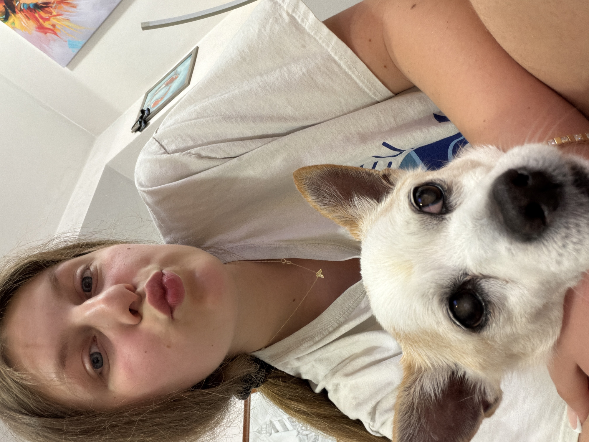 The image is a close-up, selfie-style photograph featuring a young woman and a small dog. The woman, with blonde hair tied back, is in the foreground, making a kissing face towards the camera. She is wearing a light-colored t-shirt and a delicate gold necklace with an initial pendant. A portion of the dog's face is visible in the lower part of the frame, with its head resting on or near the woman's arm. The background suggests an indoor setting with a white wall, artwork hanging on the wall, and a framed picture. There is no discernible evidence to pinpoint a specific location within Burgas, Bulgaria, or any other city. The scene is intimate and casual, capturing a moment between the woman and her pet.