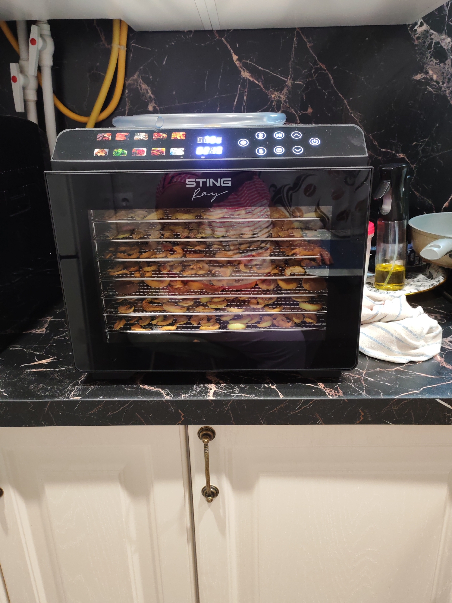 A black food dehydrator with a clear glass door sits on a black marble countertop. The dehydrator is filled with multiple trays of sliced food, likely fruit, which are uniformly arranged. The control panel at the top of the dehydrator displays digital readouts and illuminated icons, suggesting it is currently in operation. To the right of the dehydrator, a small glass bottle containing yellow liquid, possibly oil, and a white dish towel are present. The background features a black marble backsplash. The scene appears to be in a domestic kitchen environment. The location of Тешевичи, Russia, is not evident from the image.