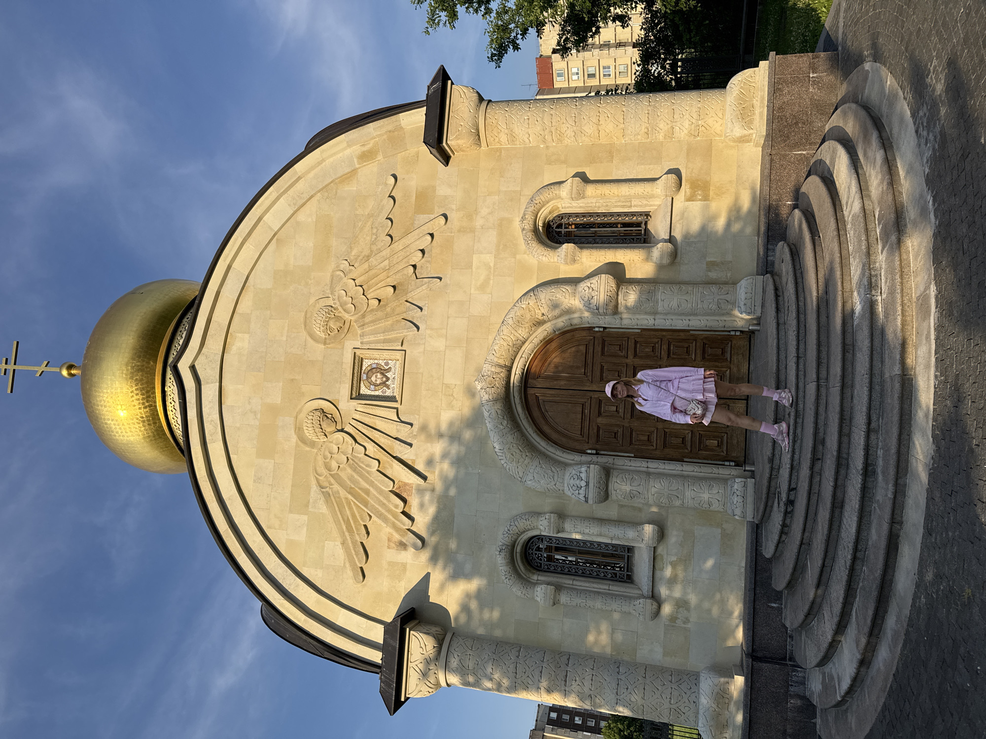 The image shows a woman standing on the steps of a church with a golden dome. The church is made of light-colored stone and has intricate carvings. The woman is wearing a pink outfit and a pink hat. The sky is blue with some clouds. There are trees and buildings in the background. The church appears to be the Church of the Assumption of the Blessed Virgin Mary in Kirishi, Russia.