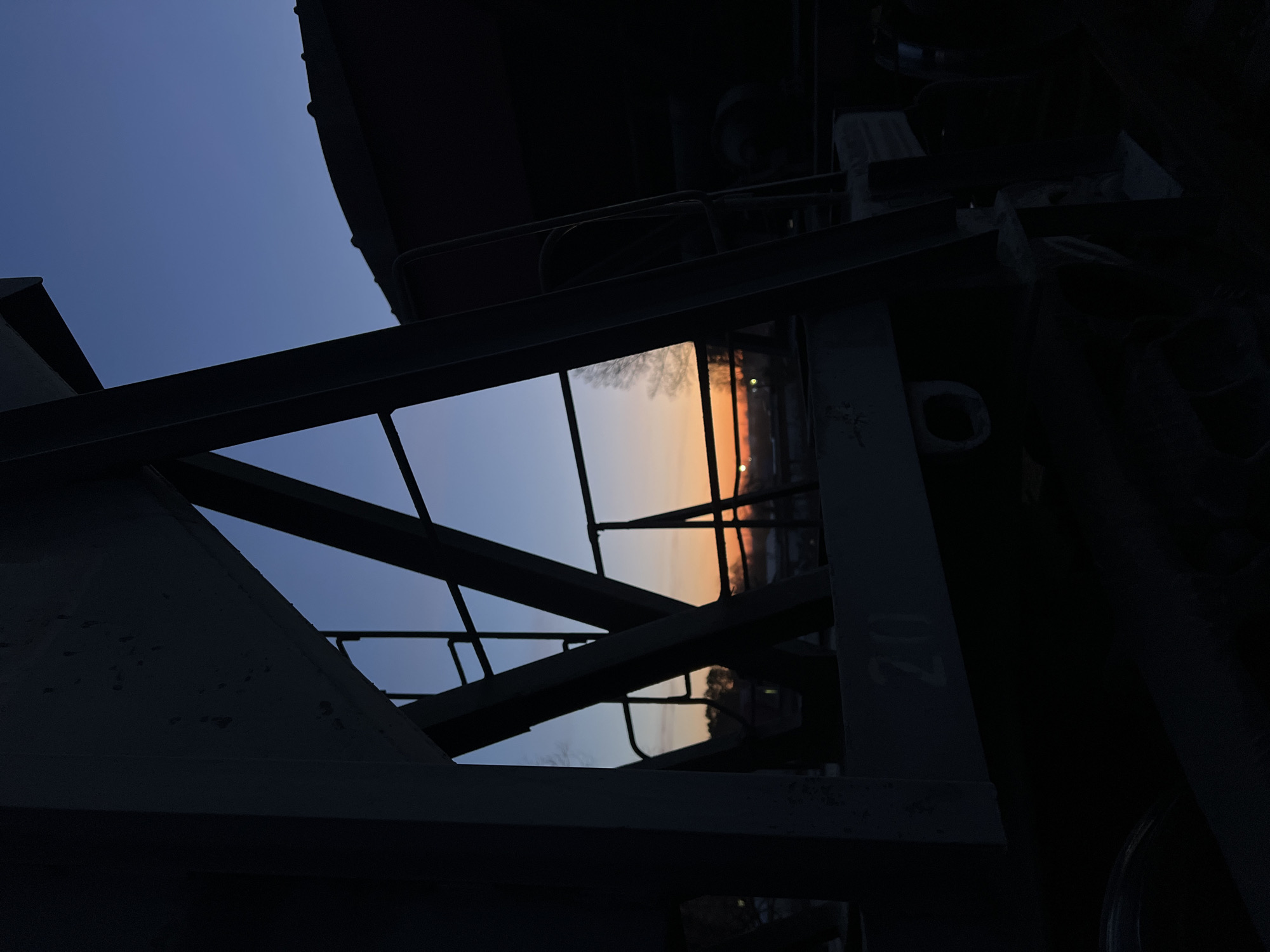 The image captures an outdoor scene in Chaikovsky, Russia, at twilight, showcasing a clear sky transitioning from deep blue to warm orange and peach tones, indicative of either sunrise or sunset. The foreground and midground are dominated by the dark, silhouetted framework of heavy metal beams and girders, possibly part of an industrial structure, bridge, or building. These elements form sharp, geometric lines and create significant areas of shadow within the composition.
Through a large, irregular opening within this dark, angular framework, the colorful sky is prominently visible. Reflections of the sky and faint outlines of bare tree branches or distant structures are discernible on what appears to be a reflective surface or window embedded within the metal construction. There are no people visible, and no overt activity is taking place; the scene is static and serene. The overall impression is one of stark beauty, juxtaposing rigid man-made structures with the soft, ephemeral colors of the natural sky. On one of the dark metal beams towards the right side, the number "02" is faintly visible.