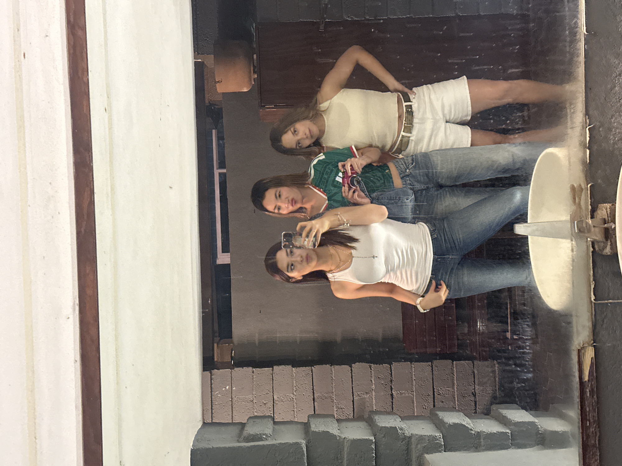In Puebla, Mexico, three young women pose for a mirror selfie. The woman on the left wears a white tank top and blue jeans, with a cross necklace. The middle woman sports a green and white sports jersey over denim overalls and holds a pink camera. The woman on the right is in a light-colored tank top and white shorts with a beige belt. They are in a dimly lit room with a brick wall to the left and a dark, possibly wooden, structure to the right. A sink is visible in the foreground, suggesting the reflection is in a bathroom or washroom. The overall ambiance is casual and youthful.
