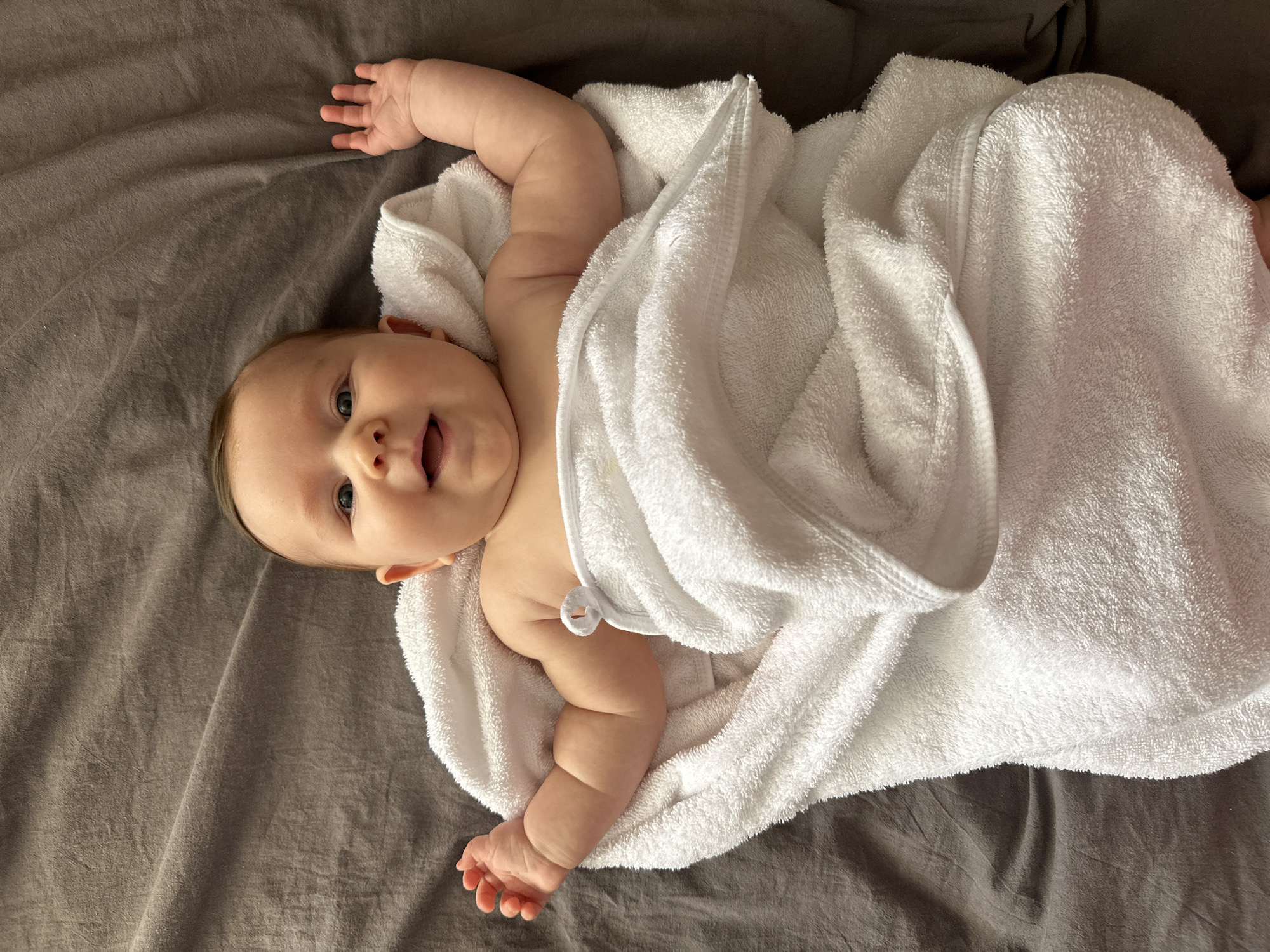 A baby, appearing to be an infant, is lying on a dark grey, textured blanket or sheet, possibly on a bed. The baby is partially wrapped in a thick, white, fluffy towel, which seems to be a hooded towel with a small white loop detail visible near its neck.
The baby is looking directly at the camera with an open-mouthed, smiling or curious expression. Its eyes are wide and dark, and it has fair skin with visible chubby cheeks. Its head is positioned towards the upper left of the frame. Both of the baby's arms are outstretched above its head, with its tiny hands open. One leg and foot are also visible, peeking out from under the white towel on the lower right side.
The lighting is soft and natural, suggesting an indoor setting during daylight hours. The overall scene conveys a feeling of comfort and contentment, perhaps after a bath. There is no visible text in the image. The provided location context is Chaikovsky, Russia.