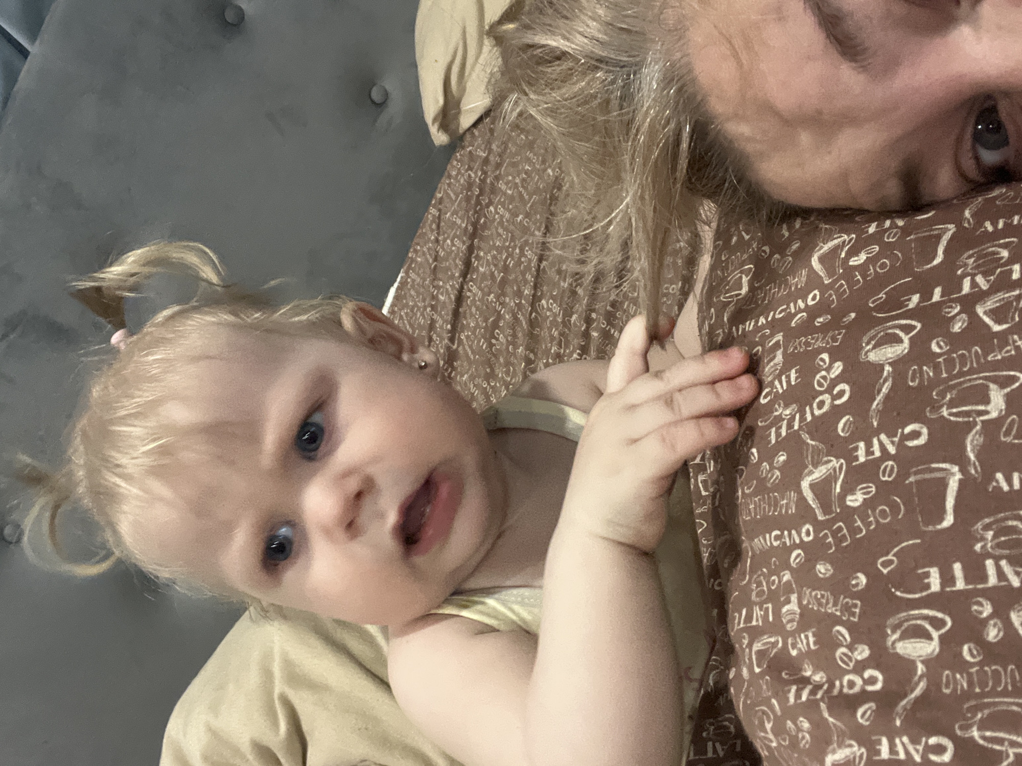 A fair-skinned baby with blonde hair in pigtails is lying down next to an adult. The adult has blonde hair, and only the adult's forehead and eye are visible in the upper-right corner of the image. The baby is looking at the camera with an open mouth. The baby is wearing a light-yellow top. The adult is wearing a brown shirt with a white coffee-themed pattern. The baby and adult are on bedding. Behind the baby is a light-tan pillow, and to the left is a gray, buttoned headboard. The image appears to be taken indoors. There is no indication of the location being Vesyoloye, Russia.