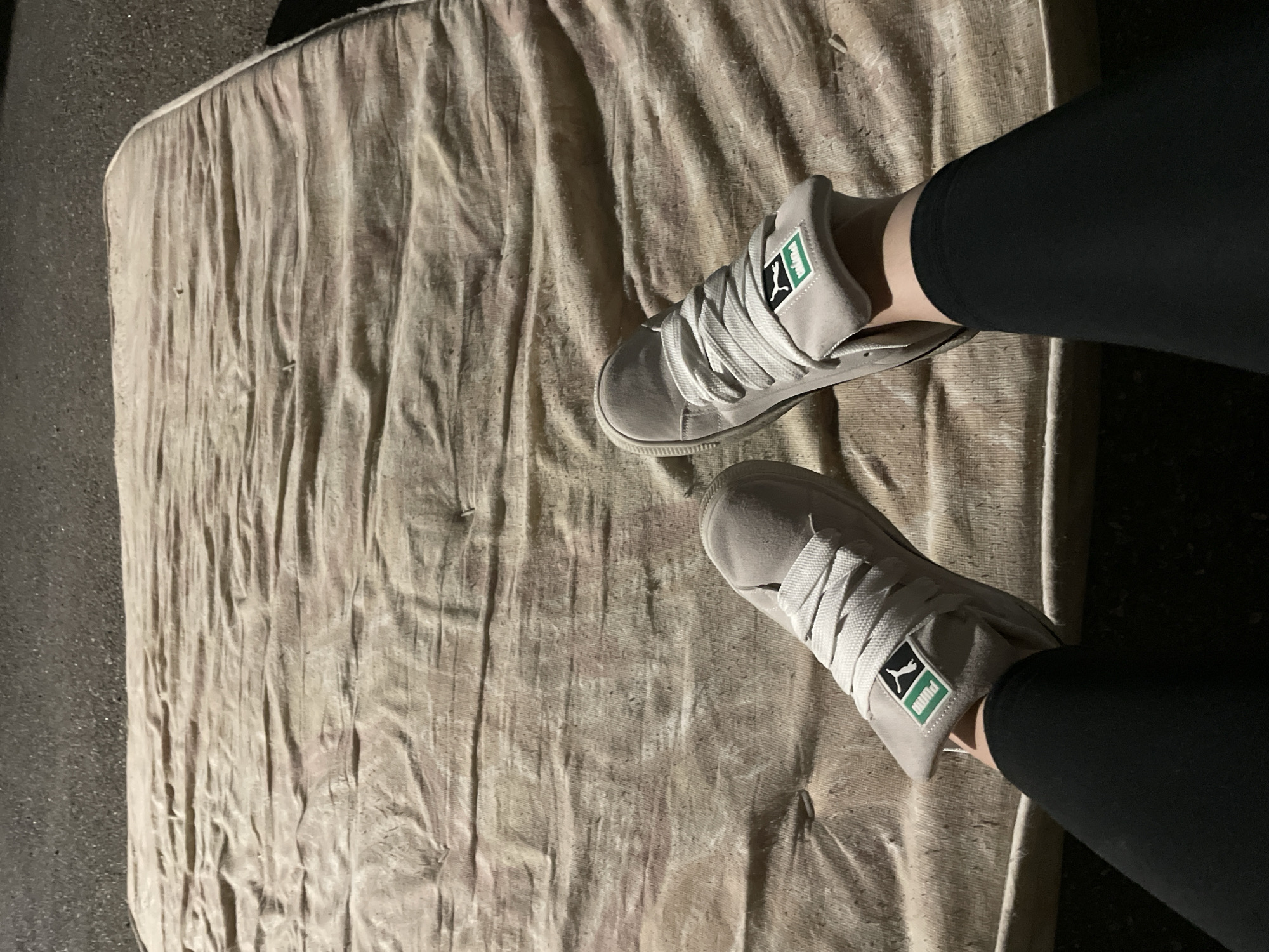 The image shows a person wearing black leggings and grey Puma sneakers standing on a dirty, possibly discarded, mattress. The mattress has a light-colored, wrinkled fabric cover. The background is dark and indistinct, suggesting an outdoor location at night. The scene is devoid of any explicit indicators of location within Yerevan, Armenia.