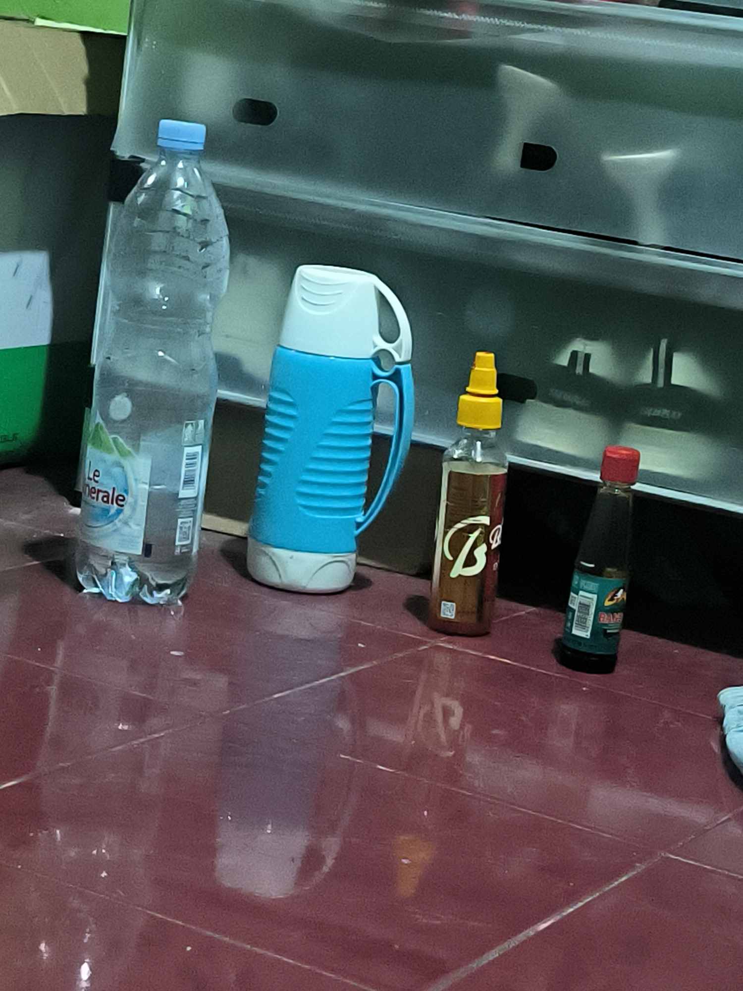 In Bumi Mulyo, Indonesia, a collection of household items rests on a red tiled floor. A large, clear plastic bottle of "LeMinerale" water stands upright, its label partially visible. To its right, a blue and white thermos sits on the floor. Further to the right, two small condiment bottles are placed side-by-side. The first, taller bottle, has a yellow cap and a label with stylized lettering. The second, shorter bottle, has a red cap and a dark label. In the background, a metallic surface with horizontal indentations is visible.