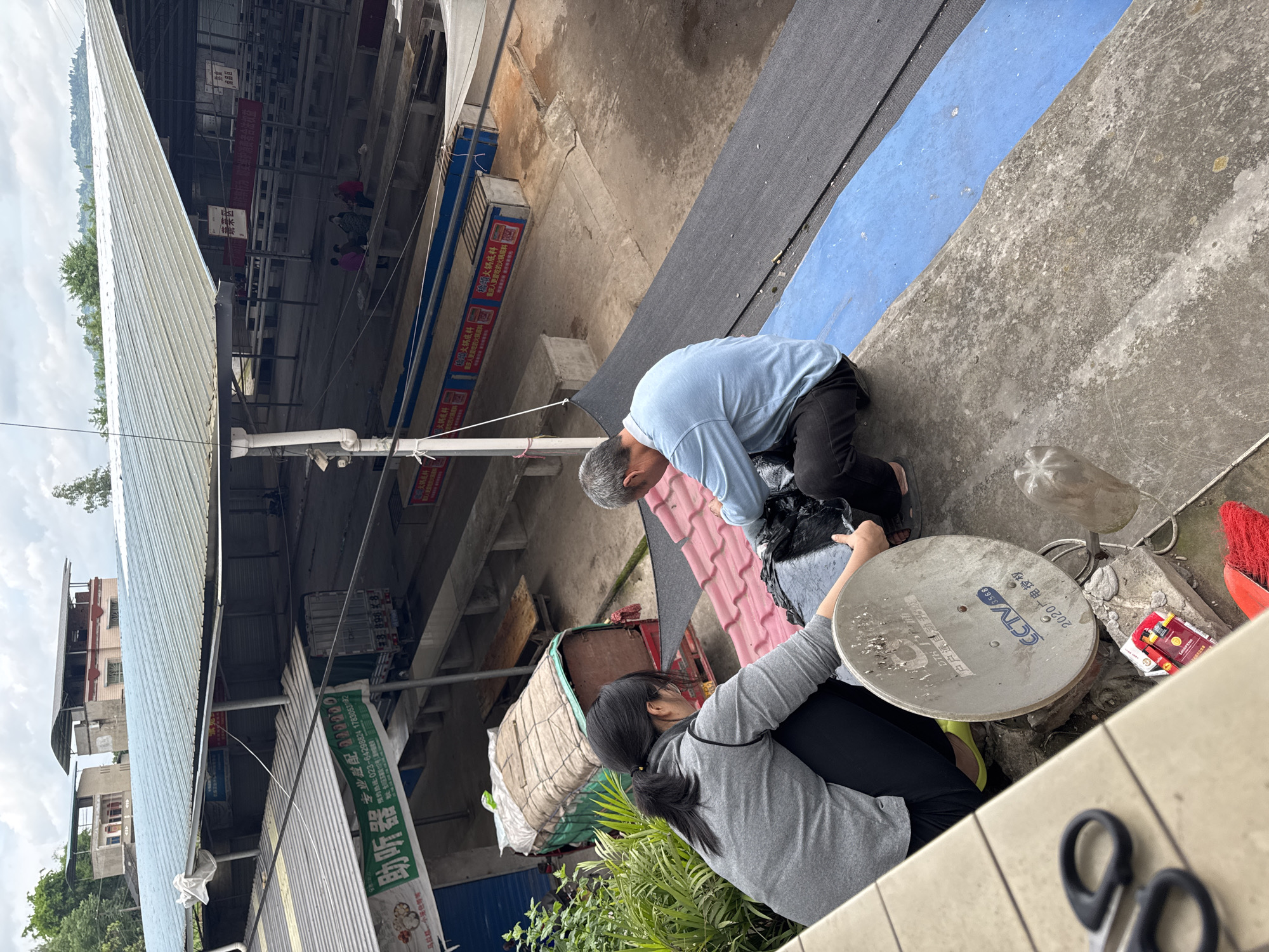 Here is a detailed description of the image:
**Setting:** The image seems to be captured in an outdoor market area in Hechuan, China. There are partially visible buildings and shops to the left with signs in Chinese.
**People:** Two individuals are visible.
- A person wearing a light blue shirt and dark pants appears to be crouching down and working on something. They seem to be older, with gray hair.
- Another person wearing a gray long-sleeved shirt and dark pants is kneeling, holding a satellite dish. This person has dark hair pulled back in a ponytail.
**Objects:**
- Satellite dish with Chinese writing on it (likely related to CCTV)
- Scissors
- Cigarette pack
- Broom
- Other unidentified items surrounding the two people
- Various shop signs and awnings in the background
- Plants visible in the lower-left corner
**Actions/Interactions:** It appears the two individuals are possibly involved in assembling or repairing the satellite dish. The older individual in the blue shirt seems to be working on something on the ground, while the person with the ponytail is holding the dish.
**Environment:** The setting is cluttered and busy, typical of a market area. The light suggests it's daytime. There are signs of wear and tear and ordinary market surroundings.