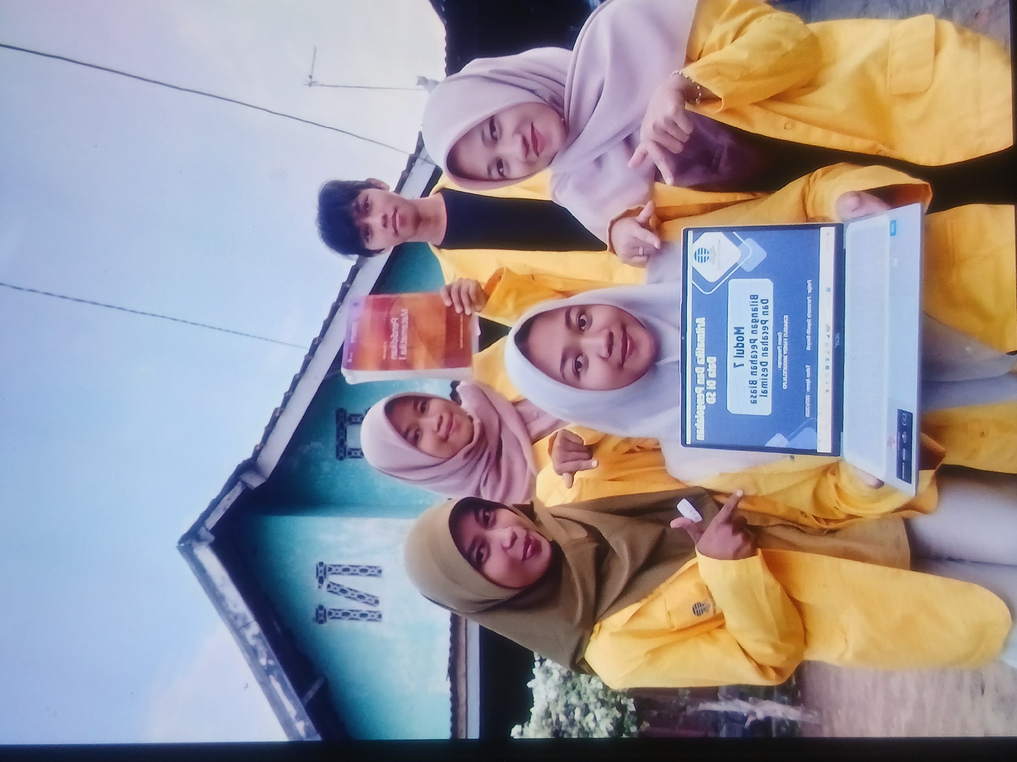 In Setia Bakti, Indonesia, a group of five young people, four women and one man, pose for a photograph outdoors. The women are all wearing hijab and yellow jackets, while the man wears a black shirt and a yellow jacket. The group is gathered in front of a light blue house with a dark roof and a decorative "M" on the facade. One of the women is holding a book with a colorful cover, and another is holding a laptop. The people appear to be in good spirits, with some smiling and others making playful gestures. The image is taken from a low angle, and the lighting suggests it was taken during the day. The background shows some vegetation and a utility pole with wires.