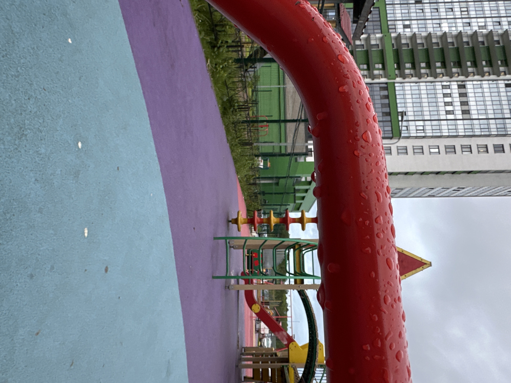 The image shows a children's playground in Kudrovo, Russia. A curved red metal bar covered in water droplets dominates the foreground, partially obscuring the view. The playground surface is a mix of light blue and purple sections. In the background, partially visible through the red bar, is a playground structure with green metal framing and yellow and red components. A tall residential building with a grid-like pattern of windows, white walls, and green accents stands in the distance. Beyond the playground, there's green foliage and a metal fence. The sky appears overcast.