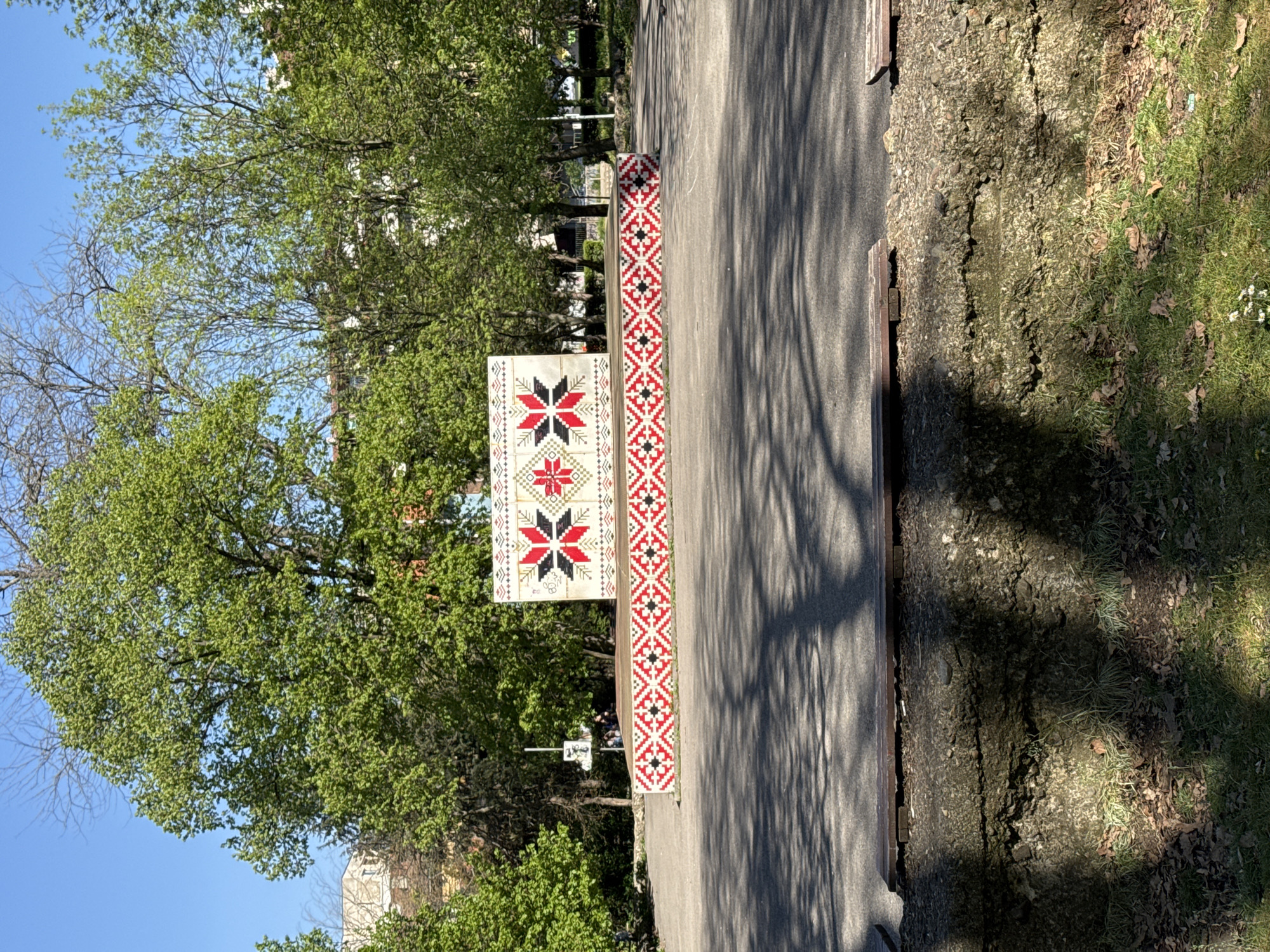 The image shows a street scene in Sofia, Bulgaria, featuring decorative concrete benches along a pedestrian walkway. The benches are adorned with traditional Bulgarian geometric patterns in red, black, and white. The walkway is paved with asphalt and lined with trees providing shade. On the left, a tree is visible with green foliage, against a clear blue sky. The adjacent ground has grass and some weeds. A building is visible in the distance.