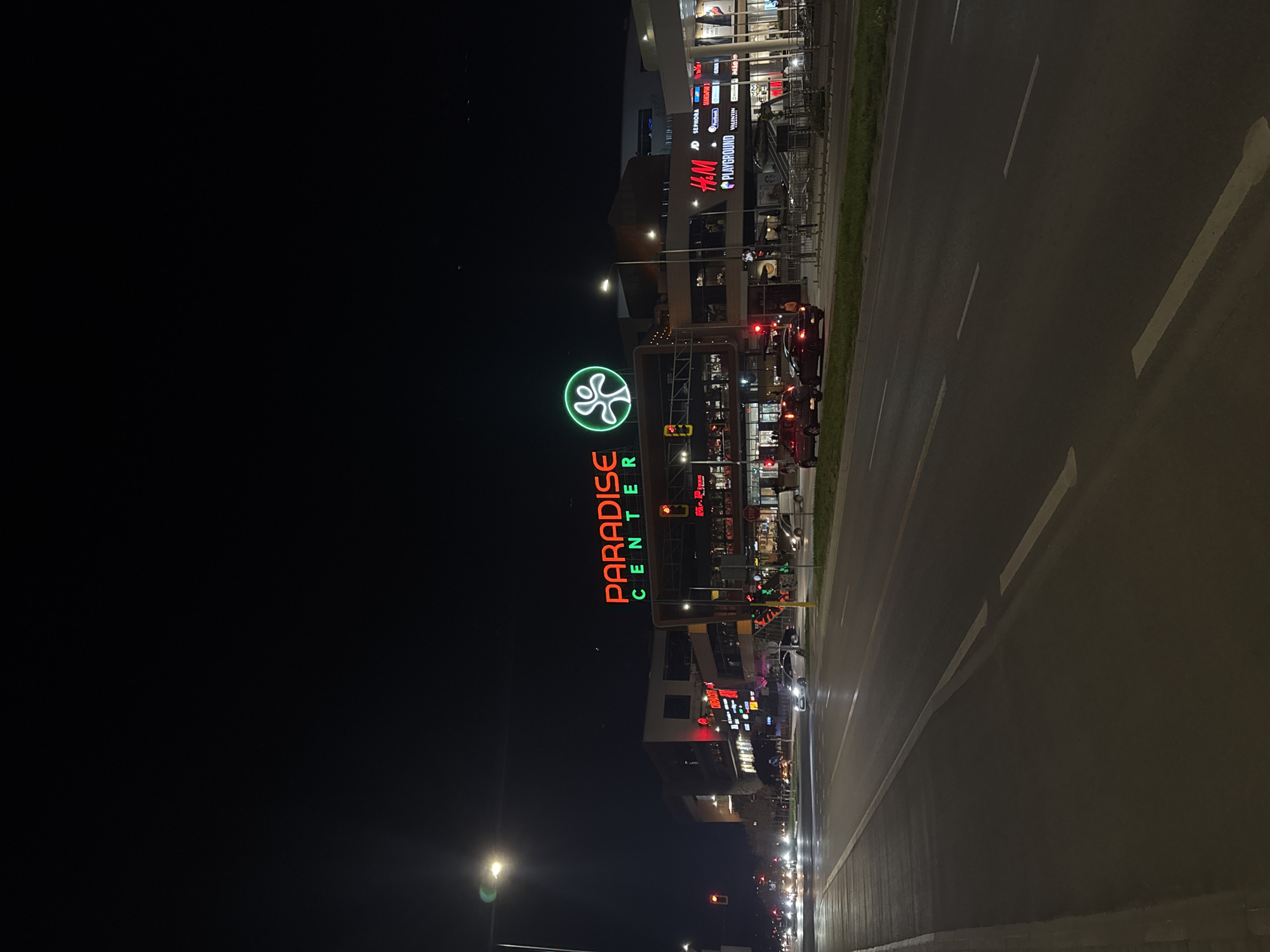 At night, Paradise Center, a shopping mall located in Sofia, Bulgaria, is visible with its illuminated logo and signage. The mall features retail outlets such as H&M. A road with lane markings runs alongside the mall, and multiple cars are waiting at a traffic light. Streetlights illuminate the surrounding area. The sky is dark.