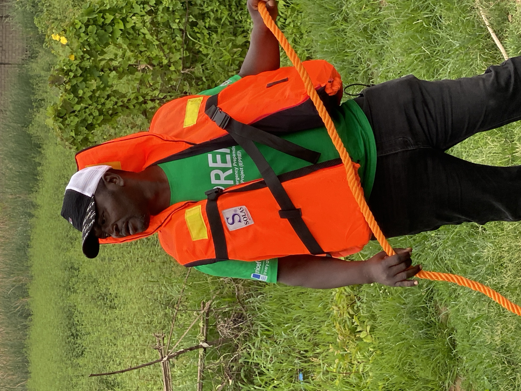 A Black man wearing a green t-shirt, an orange life vest, and a black and white baseball cap stands outdoors in a grassy area, holding an orange rope. The man is looking to his right. The t-shirt has white text that appears to read "Emergency Preparedness Project (EPREP)". The life vest has two yellow reflective strips and a circular logo with an "S" on it. The background consists of dense green vegetation and some yellow flowers. The scene is likely located in the city of Kawo, Nigeria, given the context of the surrounding environment and potential organizational affiliations.