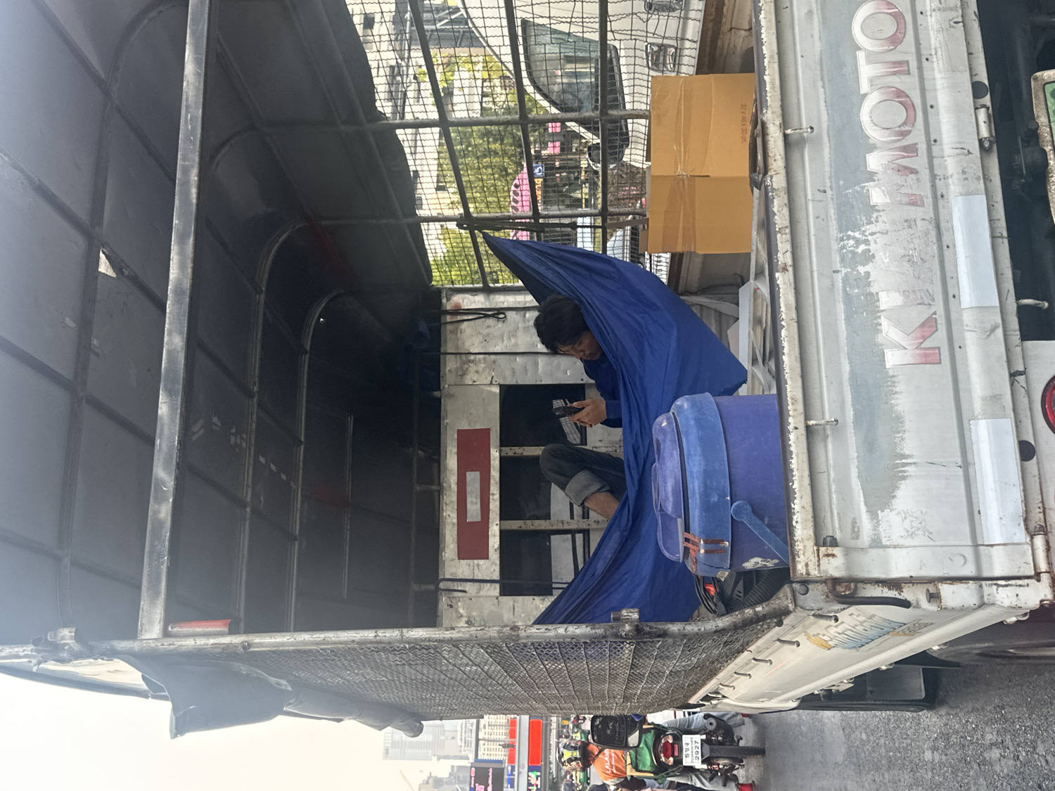 The image presents a view into the back of a weathered white utility truck, likely parked on a busy street in Bangkok, Thailand. Inside the truck, a man is seen resting or waiting, partially enveloped by a bright blue tarp or fabric. He is wearing a dark shirt and jeans, engrossed in looking at a smartphone held in his hands. Beside him is a large blue cooler or plastic container, and a cardboard box marked with "3D-46L x 30L". The truck's interior features a metal frame and various reflective markers, including a red rectangular one with the letter "D".
The truck's exterior side panel prominently displays the brand "KIMOTO" in large silver and red lettering, with a smaller "K" logo below it. The scene outside suggests daytime with clear, bright lighting. The background reveals an urban street teeming with motorcycles, many appearing to be delivery bikes due to their attached storage boxes. One scooter's license plate is partially visible, showing Thai script and the numbers "593 2927". Buildings and overhead infrastructure line the distant street, completing the bustling city environment. The man appears calm and focused on his device, suggesting a moment of downtime amidst the active surroundings.