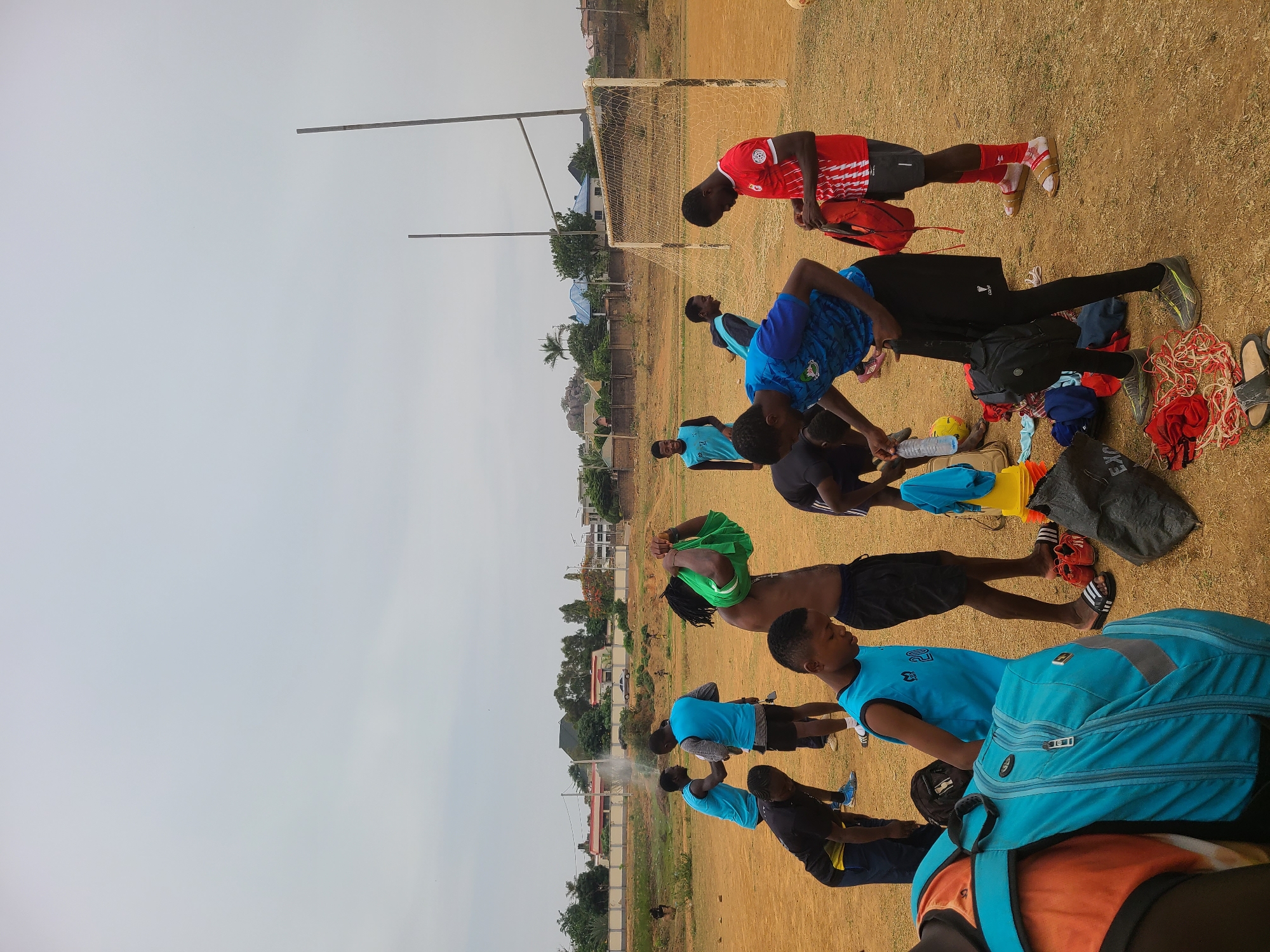 In Barikin Jirga Summa, Nigeria, a group of young men, presumed to be soccer players, are outdoors on a field. They are stretching or bending over, possibly as part of a pre-game warm-up or post-game cool-down routine. Various bags, shoes, clothing items, and a water bottle are scattered on the ground near the players. A soccer goal net stands in the background. The sky is overcast. Buildings and trees are visible in the distant background.
