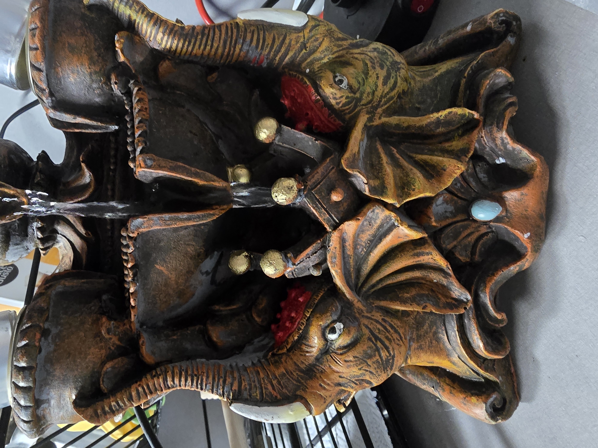 A close-up, eye-level, indoor shot shows a decorative, multi-tiered water fountain shaped like elephants. The fountain is made of a dark brown and orange material, with carved details resembling the texture of elephant skin. Two elephant heads face forward, with large ears and tusks. The top tier features a small cascade of water flowing down. Small gold orbs are embedded in the fountain's structure, and red accents adorn the elephants' foreheads. A light blue teardrop-shaped gem is positioned at the base of the fountain. The background is blurred and shows shelves with various objects, including what appears to be a bottle with a label. The fountain sits on a grey surface, possibly a table or countertop. The scene is brightly lit, emphasizing the textures and colors of the fountain.