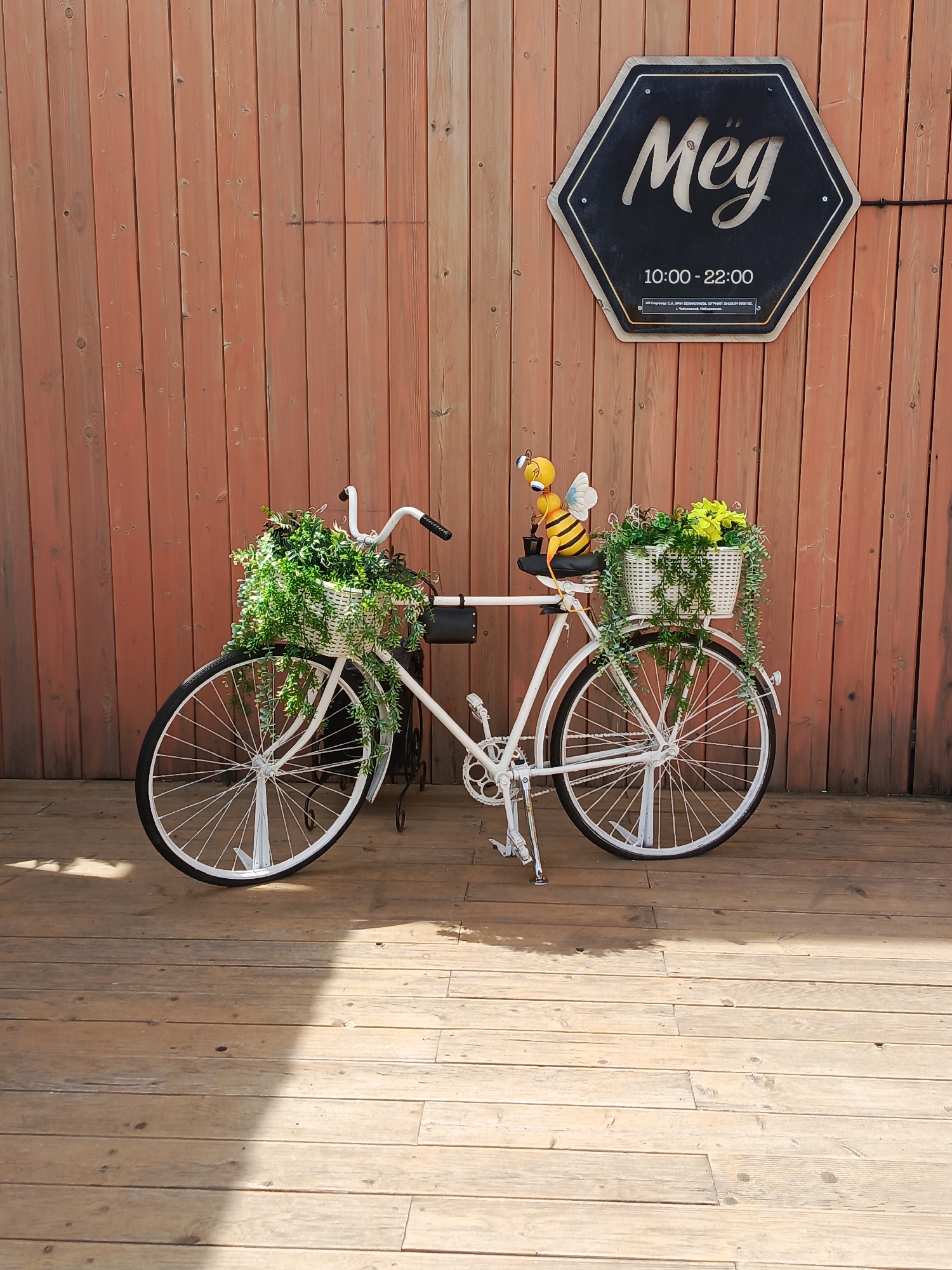 A white bicycle with baskets filled with greenery and a bee doll is parked in front of a wooden wall with a sign that reads "Meg" and "10:00-22:00" in Chaikovsky, Russia. The bicycle is on a wooden deck.