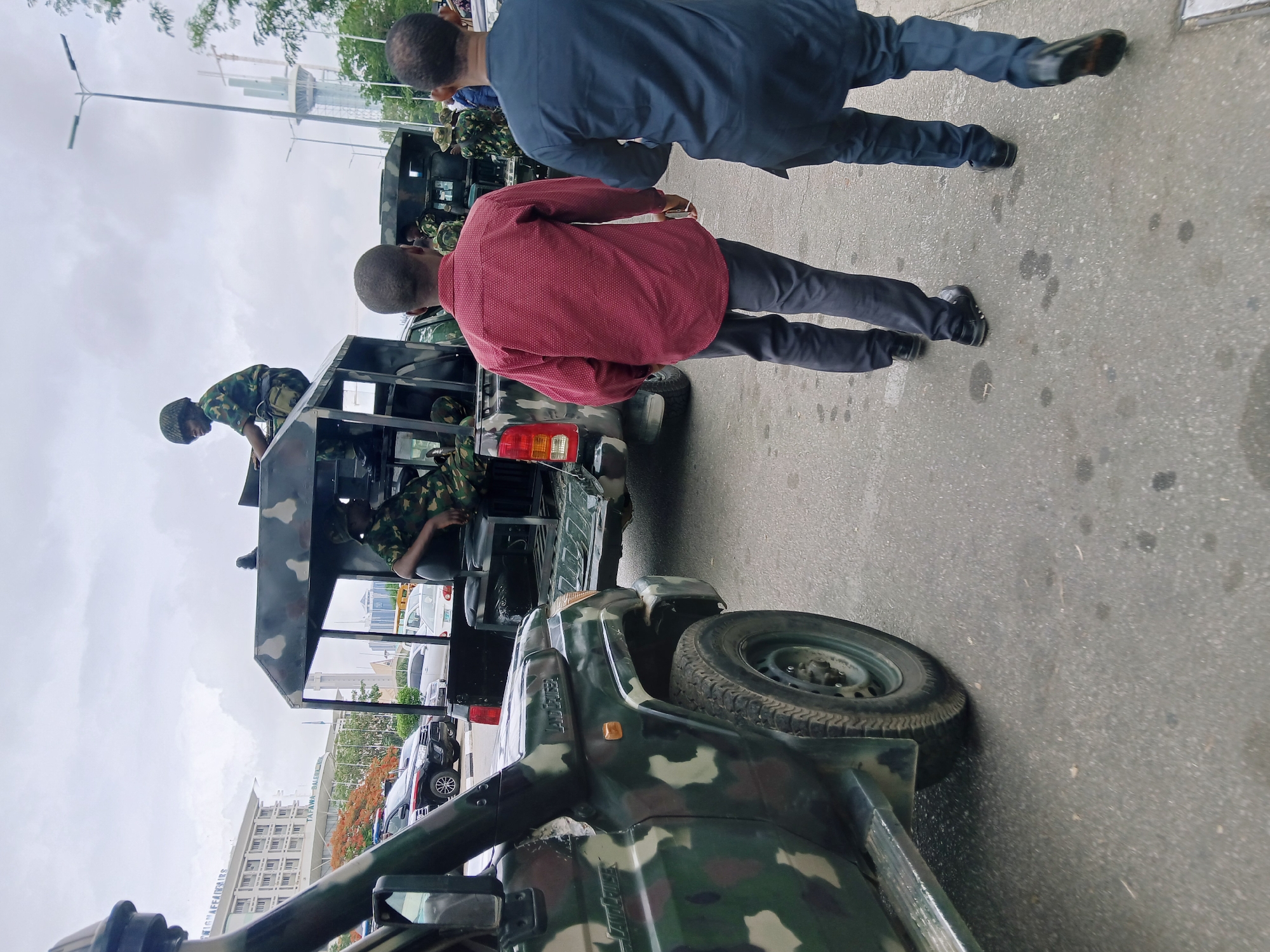The image depicts a street scene in Abuja, Nigeria, featuring two men walking past a military vehicle. The vehicle is a camouflage-painted truck with soldiers in military fatigues visible in the back. One soldier is standing on the back, while another is seated. The two men walking past are dressed in civilian clothes: one in a blue shirt and dark pants, and the other in a maroon shirt and dark pants. The street is paved, and buildings are visible in the background, including the Tafawa Balewa Building. The sky is overcast.