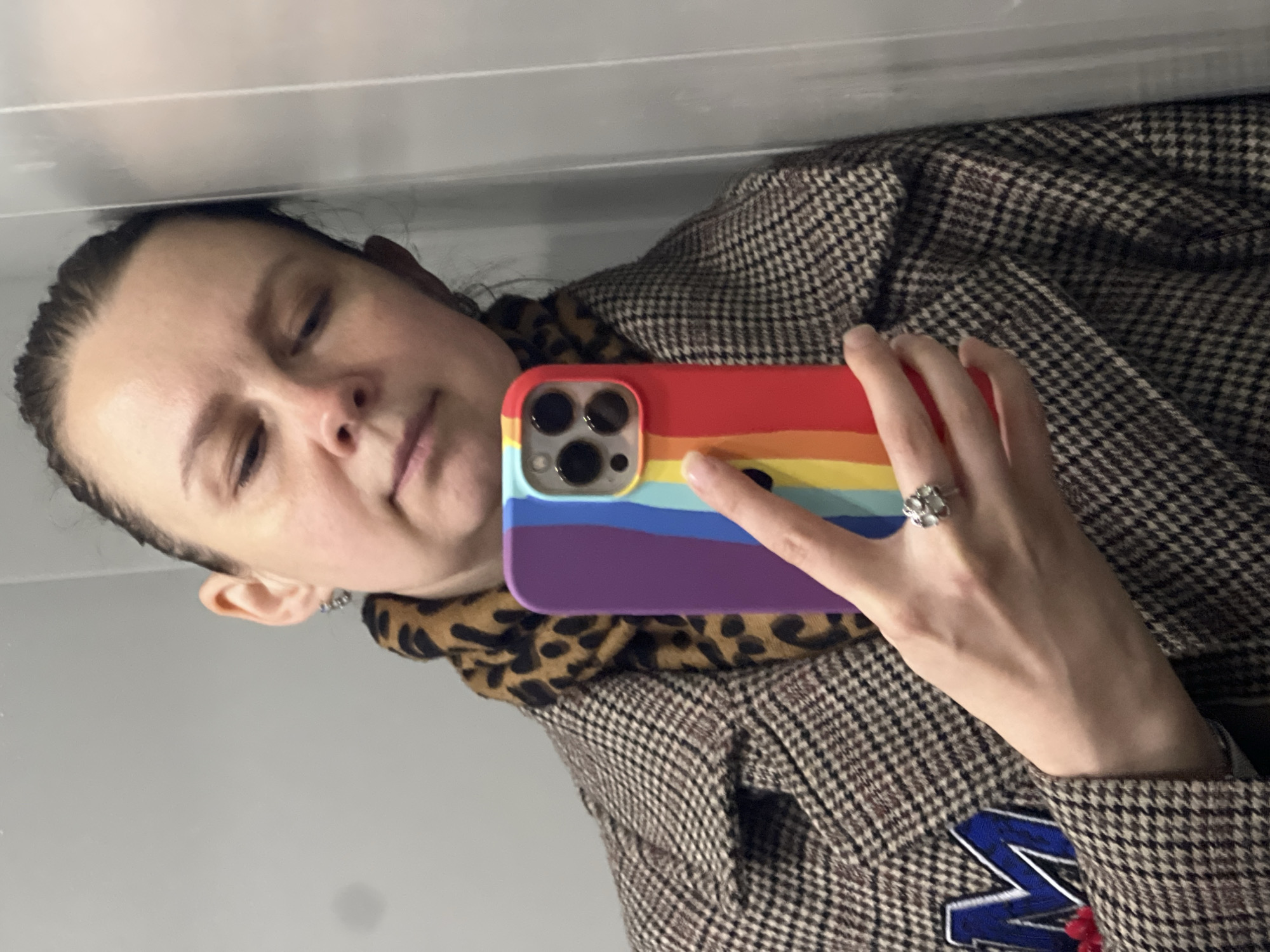 A woman is taking a selfie indoors. She is wearing a brown and white houndstooth patterned coat with a patch depicting a blue "M" and a red detail. She has a leopard print scarf or top underneath. She is holding a smartphone with a rainbow-colored case with its camera pointed at her face. She is wearing a ring on her ring finger. The background is a light grey wall. The exact location within Chaikovsky, Russia, is undeterminable from the image alone.