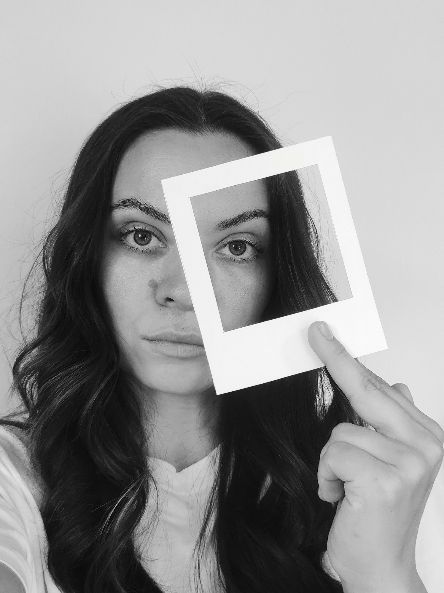 A black and white close-up shot shows a woman holding a white, square, picture frame in front of her face. The frame covers the right side of her face, from forehead to chin, with a finger holding it in place. Her left eye and the left side of her face are visible. She has dark, wavy hair. Her expression appears serious. The background is a plain, light-colored wall. The location is unknown.