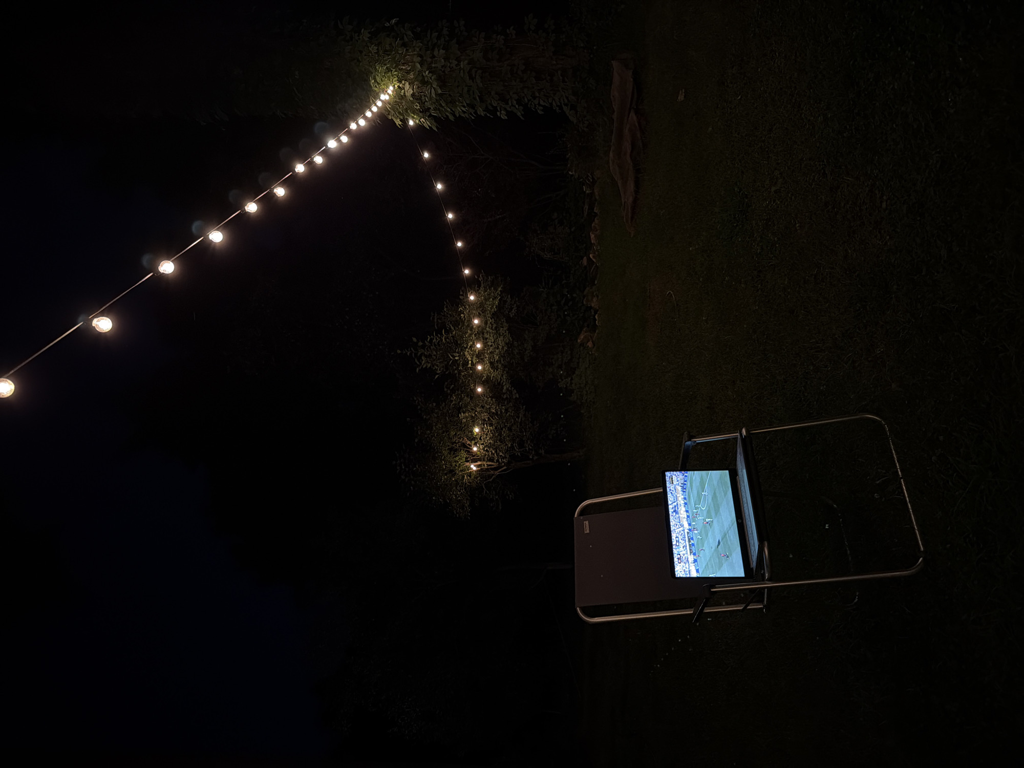 An outdoor scene at night in Udelnaya, Russia. A folding chair sits on a grassy lawn. A laptop computer is open on the chair, displaying a soccer match. String lights are visible in the background, illuminating trees and the dark sky. The overall impression is one of peaceful evening entertainment.