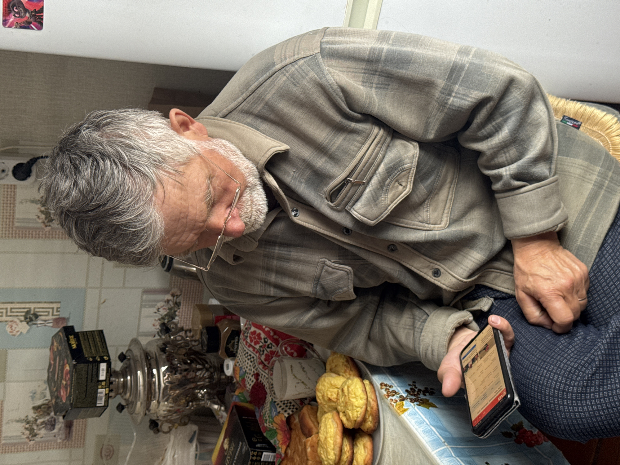 An elderly man with gray hair and glasses is seated at a table. He wears a plaid shirt and blue patterned pants. In his right hand, he holds a smartphone. On the table, there is a plate of pastries, a samovar, boxes, and a tablecloth with various patterns. The location is a room, potentially a home. The image does not confirm it is Вассята, Russia.