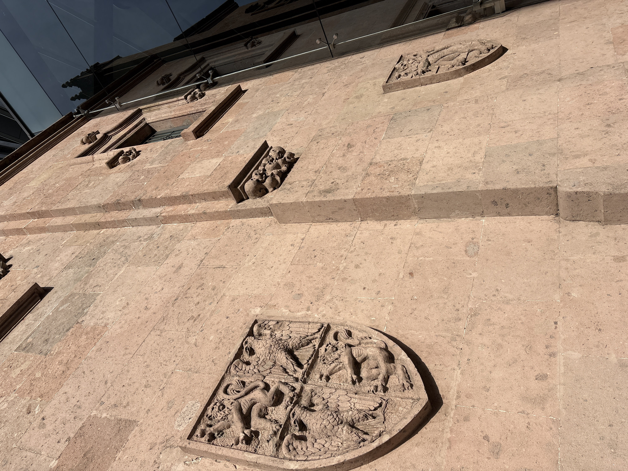 The image shows a facade of a building in Mexico City, Mexico. The building is constructed of light brown stone and features relief sculptures embedded in the walls. A large, shield-shaped sculpture depicts four heraldic creatures, with a dragon, eagle, a lion, and a griffon. Other sculptures include what appear to be human or animal figures. The upper portion of the building is partially obscured by a modern glass structure. The image is taken from a low angle. The lighting suggests a sunny day.