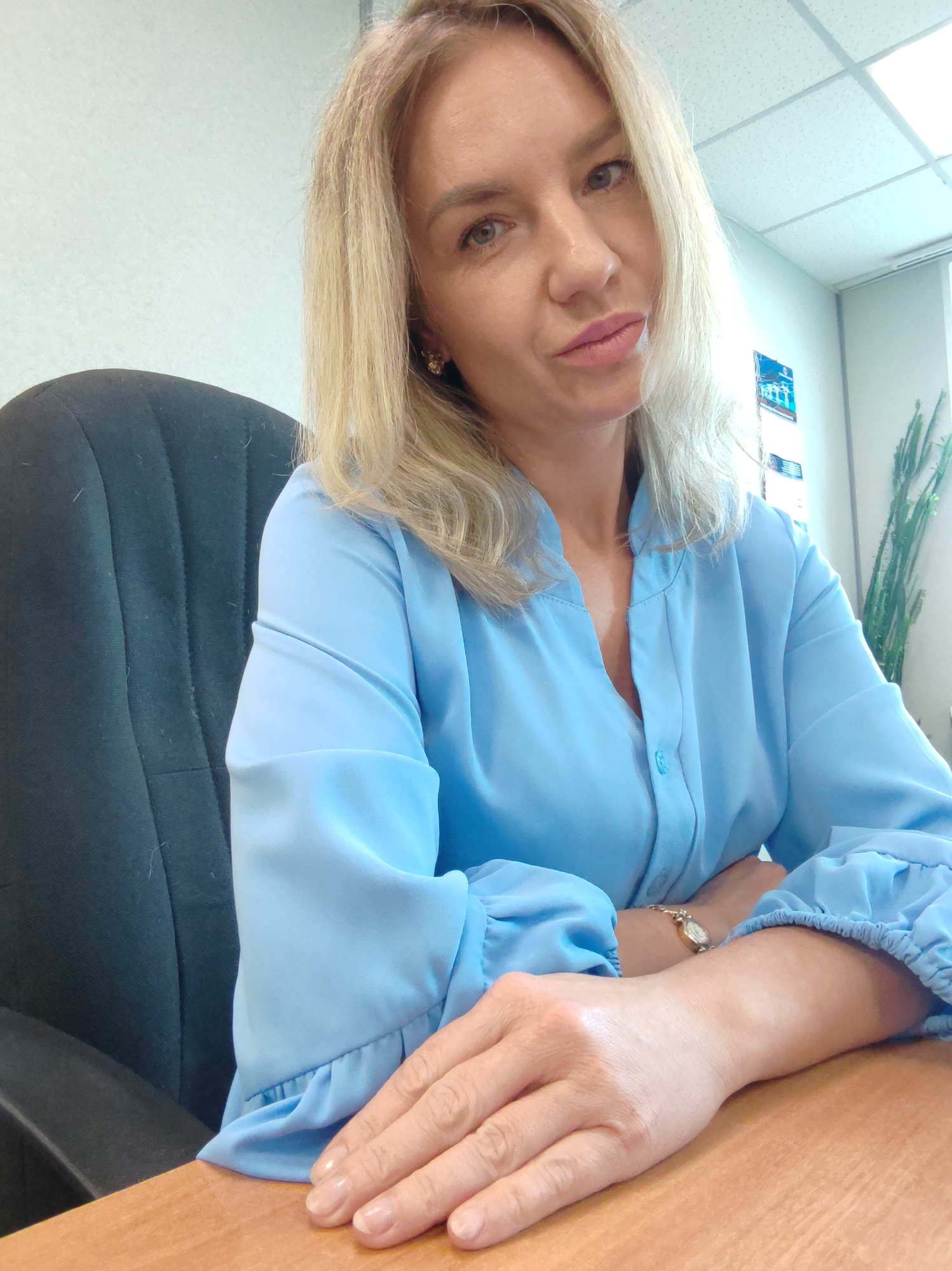 A woman with blonde hair, wearing a light blue collared shirt, sits at a wooden desk in an office. Her arms are crossed in front of her, and she is looking towards the camera with a slightly pursed-lip expression. A watch with a gold band and a round face is visible on her left wrist. The background shows a wall, ceiling lights, and a tall green plant in a pot. There are some papers or posters on the wall behind her. The lighting suggests it is daytime.