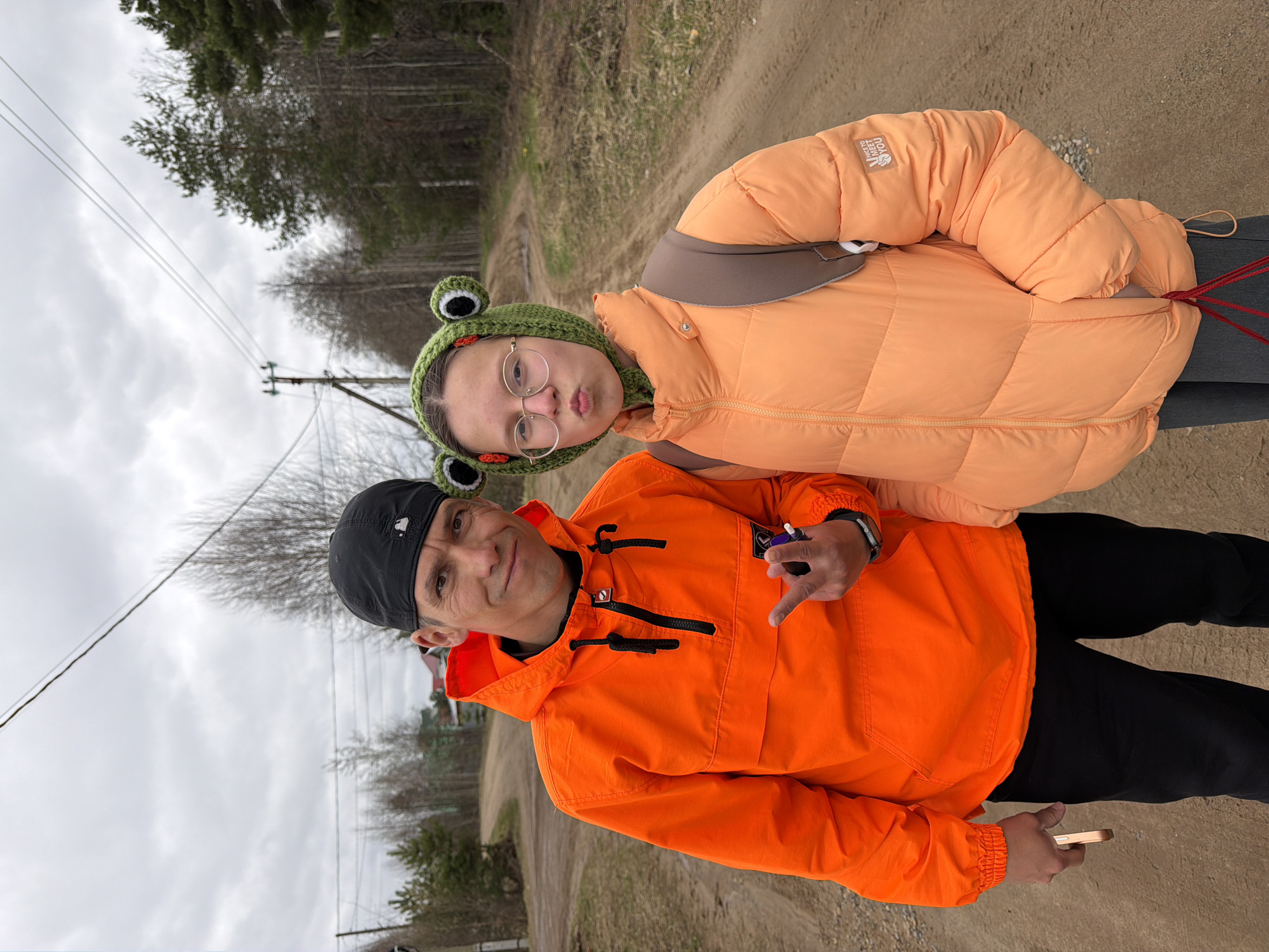 The image features two people standing on a dirt road near a wooded area, likely a rural or suburban setting. The background shows trees and utility poles, with an overcast sky above.
The person in the foreground is wearing a black baseball cap and an orange anorak with black zipper pulls and details. They are holding a small object in one hand and a cell phone in the other, and looking at the camera.
Behind and slightly to the side of this person is a younger individual with glasses and a green frog-themed hat. They are wearing a light orange puffer jacket. Their expression is a slight pout.
The location is suspected to be in or near the city of Chaikovsky, Russia, based on contextual information not visible in the image itself.