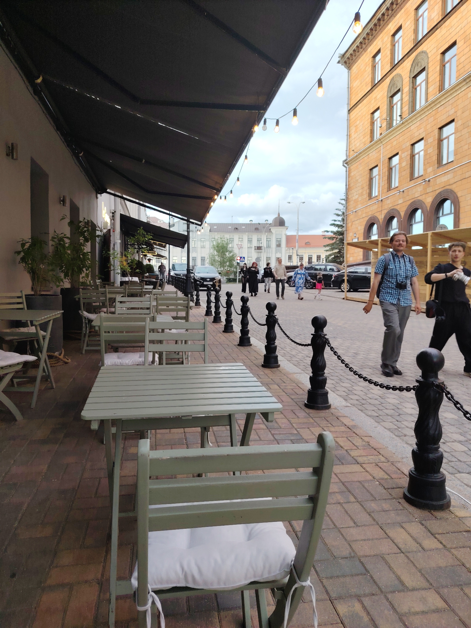 The image presents an outdoor cafe and a bustling street scene in Minsk, Belarus, under an overcast sky. In the immediate foreground and extending along the left, a series of light green wooden tables and chairs, each fitted with a white cushion, are arranged parallel to a light-colored building facade. A dark, retractable awning covers this seating area, with a string of exposed-bulb lights suspended underneath. Large potted plants line the building wall, and a coiled rope is visible on the reddish-brown brick pavement beneath the cafe tables.
To the right of the cafe, a distinct paved pedestrian area, featuring multi-toned rectangular bricks, is demarcated by a line of black decorative bollards interconnected by chains. Two men are walking in the midground: the man on the left wears a blue and white checkered shirt, gray pants, a backpack, and has a camera suspended around his neck; the man on the right wears a black long-sleeved shirt and black pants, carrying a black shoulder bag.
Further down the street, additional pedestrians include a woman in a blue patterned dress walking with a small child in a pink dress, and a man in a light jacket. Several dark sedans are parked along the street on the left. On the right side of the street, a large, multi-story building with orange-yellow brickwork features multiple rectangular and arched windows, indicating an older architectural style. In the distant background, a red traffic light, various buildings (one with a dark, domed roof), and sparse foliage are visible beneath a uniformly cloudy sky.