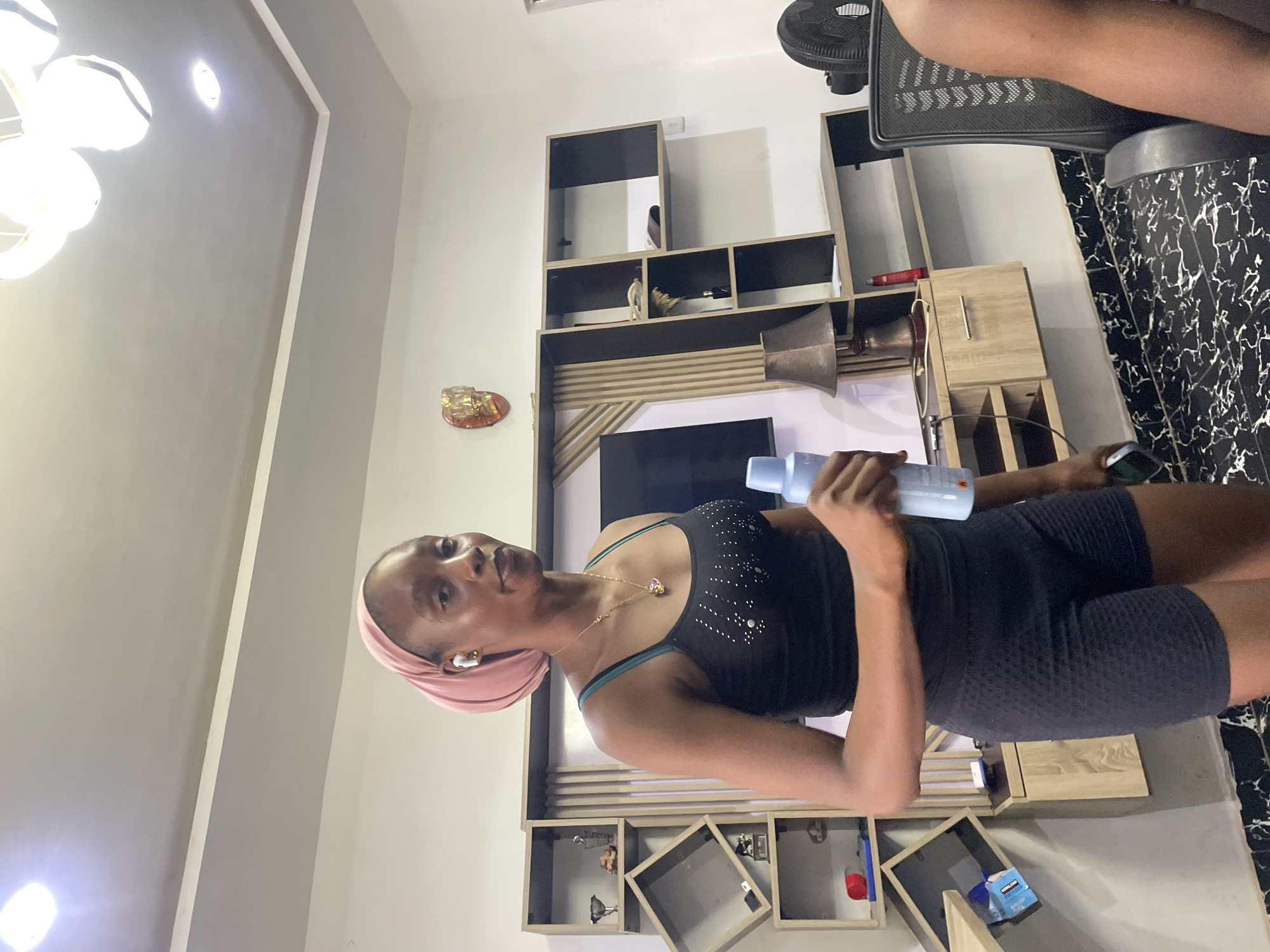 A young Black woman wearing a pink headscarf, a black tank top adorned with white rhinestones, and black athletic shorts stands in a modern living room, holding a light blue spray bottle. She has a gold necklace with a colorful pendant and is wearing white wireless earbuds. The room features a large, wall-mounted entertainment unit with shelves displaying various items, a television, and a desk with a lamp. The floor is tiled with a black and white marble pattern. The lighting suggests it is daytime, with a chandelier visible on the ceiling. The overall impression is of a well-decorated, contemporary living space.