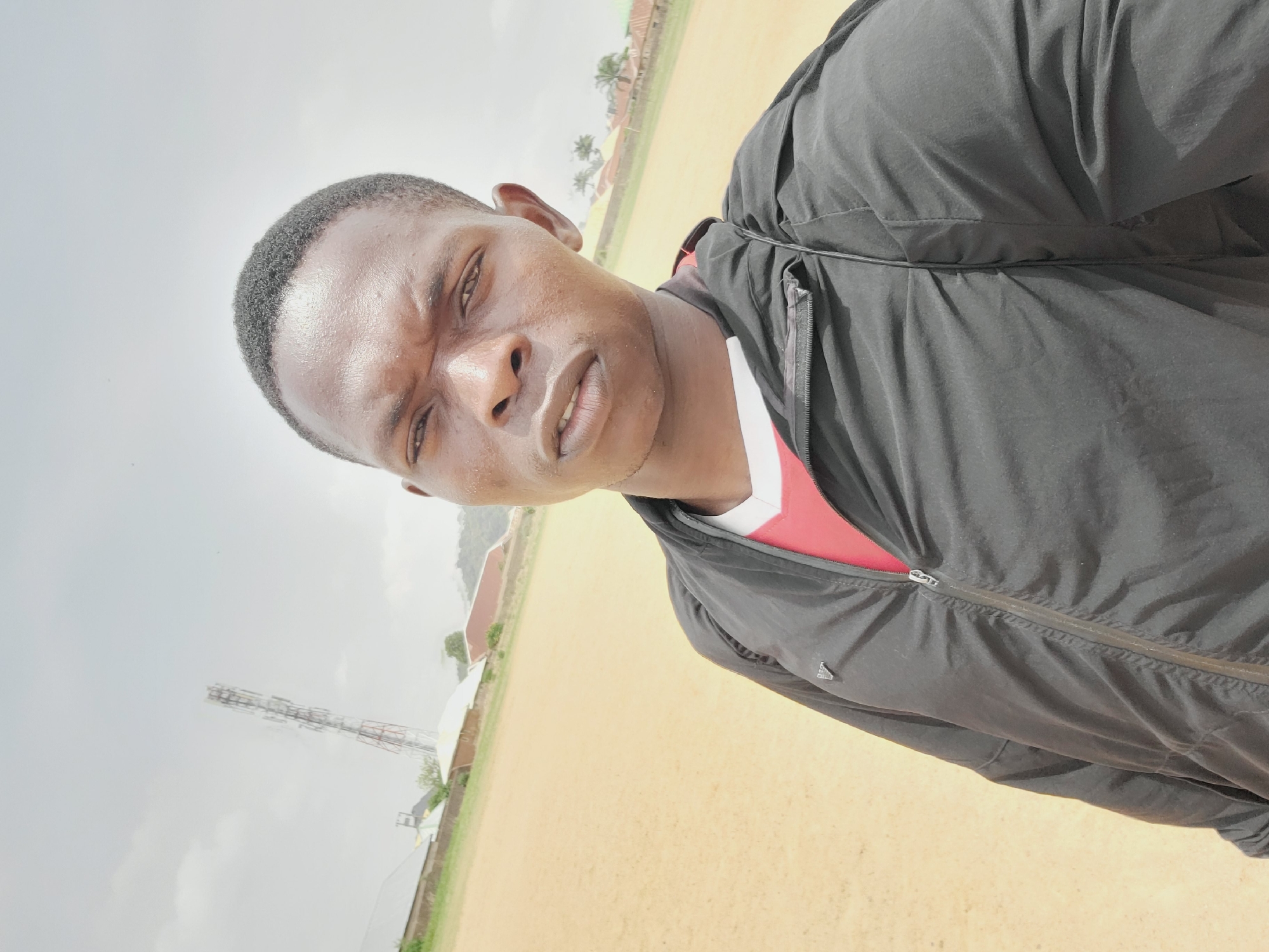 The image is a medium close-up selfie of a Black man with short dark hair wearing a black jacket over a red and white shirt. The background features a sandy field and a cloudy sky. In the distance, there are buildings, some greenery, and a tall metal communication tower. Considering the location, the setting appears to be in a rural or semi-urban area within Bukuru, Nigeria, perhaps a sports field or open space on the outskirts of the city. The man's expression is neutral or slightly stern, looking directly at the camera.