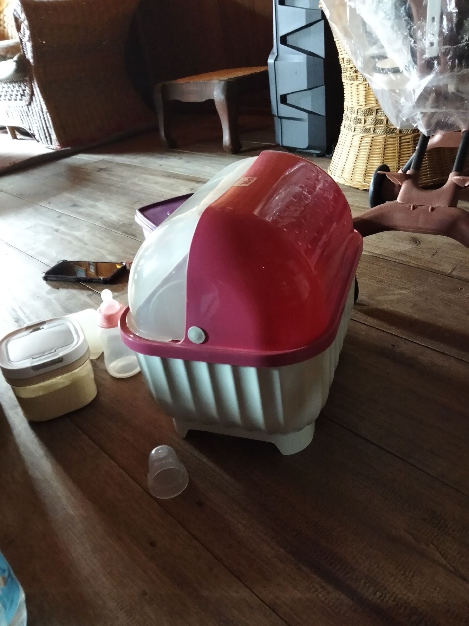 The image depicts a close-up, high-angle shot of a pink and white baby bottle sterilizer situated on a wooden floor in what appears to be a residential setting in Setia Jaya, Indonesia. The sterilizer is the central focus, with its domed pink lid and ribbed white base clearly visible. To the left of the sterilizer, a beige and white baby formula container is placed next to a clear baby bottle with a pink nipple. Further to the left, a black smartphone lies on the floor. In the background, a partially visible wicker basket and a dark wooden stool are present. The lighting suggests an indoor environment, with shadows cast by the objects onto the wooden floor. The overall scene is cluttered but suggests everyday life, possibly a nursery or a kitchen area.
