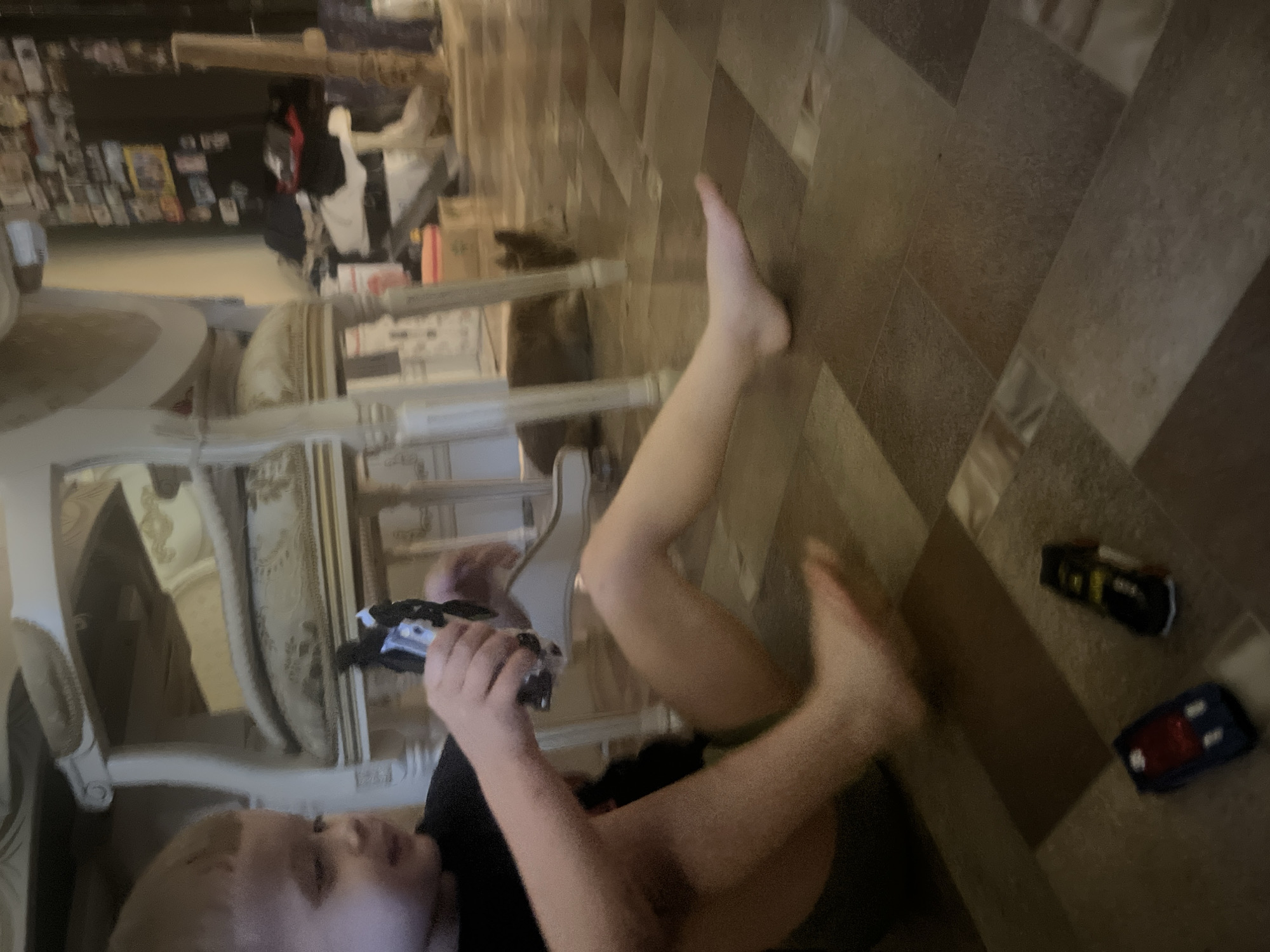 An indoor scene depicts a young child sitting on the floor, holding a toy robot. The child, with blonde hair and wearing a black t-shirt, is positioned in the foreground, their legs extended. In the middle ground, a brown cat is lying down, and a chair with ornate carvings is visible. The floor is tiled with a pattern of brown and grey squares. In the lower right corner, two toy cars, one blue and one black with yellow accents, are present. The background is blurred, showing shelves with items and framed pictures on a wall. The lighting suggests an indoor environment, likely a home.