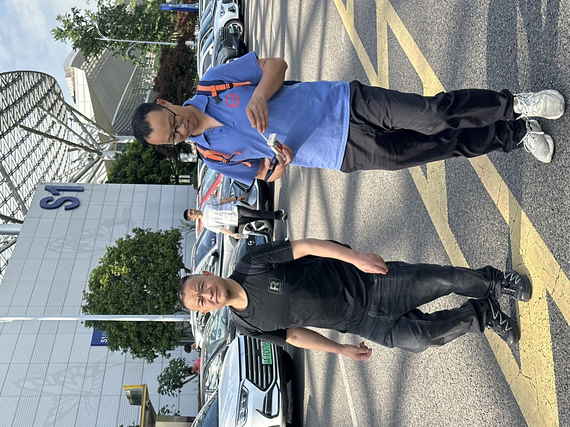 Two men stand on an asphalt surface marked with yellow crosshatch lines. One man wears a blue polo shirt, black pants, white sneakers, and glasses; he holds a pack of cigarettes. The other man wears a black t-shirt, dark denim jeans, and black sneakers. In the background are parked cars, a tree, and a building with the blue text "S1" visible. The environment suggests a location in the Liangjiang New Area, Chongqing, China.