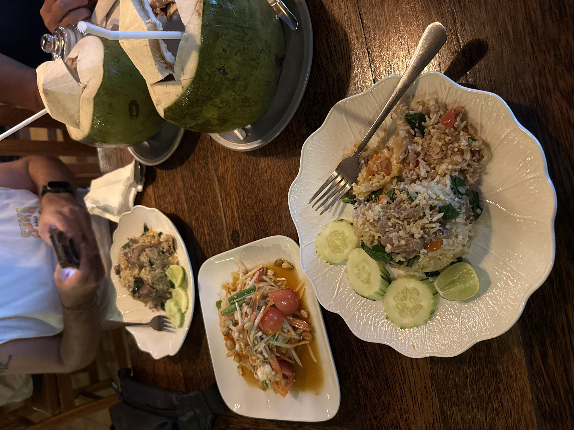 The image captures a casual evening dining scene at a restaurant or eatery in Baan Lamai, Thailand. The setting features a rustic wooden table illuminated by the flashlight of a phone, held by a person whose arm and torso (wearing a white T-shirt with partial text "SUKI" visible) are on the left. Another diner's arm is partially visible in the top left.
The table is laden with traditional Thai dishes and drinks. Two whole young coconuts, cut open and served with straws in metal stands, are prominently displayed at the top, suggesting refreshing beverages. Three plates of food are arranged on the table: a large, ruffled-edge white plate holds a generous portion of fried rice garnished with slices of cucumber and a lime wedge. A smaller, similarly shaped plate contains another serving of fried rice, also accompanied by cucumber and lime. Between these two plates, a rectangular white dish presents a vibrant papaya salad, featuring shredded papaya, cherry tomatoes, peanuts, and green beans in a savory sauce. Forks are placed on the fried rice plates, ready for dining. A black bag or pouch rests on the table below the smaller fried rice plate.
The scene suggests a relaxed and enjoyable meal, with the use of the phone's flashlight indicating a dimly lit, cozy evening atmosphere. The specific dishes – fresh coconuts, fried rice, and papaya salad – are strong indicators of a Thai culinary experience, consistent with the Baan Lamai location.