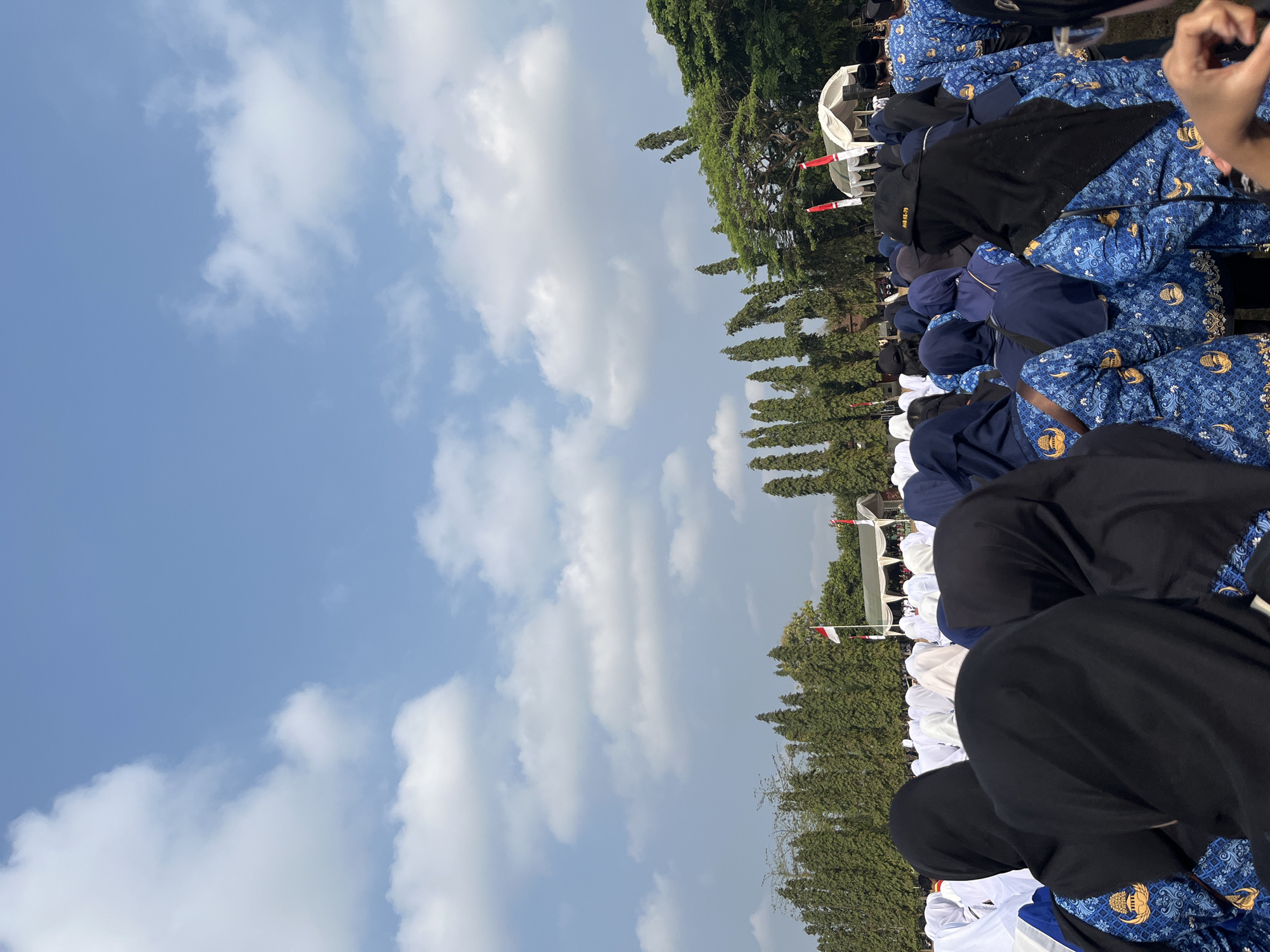 In Gembong, Indonesia, a large gathering of people, predominantly women wearing dark headscarves and blue uniforms with gold emblems, are assembled outdoors under a bright blue sky with scattered white clouds. The individuals are facing away from the camera, suggesting they are attending an event or ceremony. In the background, a line of tall, dense green trees forms a backdrop, with some taller, more sparse trees visible further back. Several Indonesian flags are displayed, one flying from a flagpole and others on smaller poles. A white tent structure is also present in the distance, indicating a potential stage or gathering area for the event. The overall scene depicts a daytime outdoor gathering in a semi-rural or park-like setting.