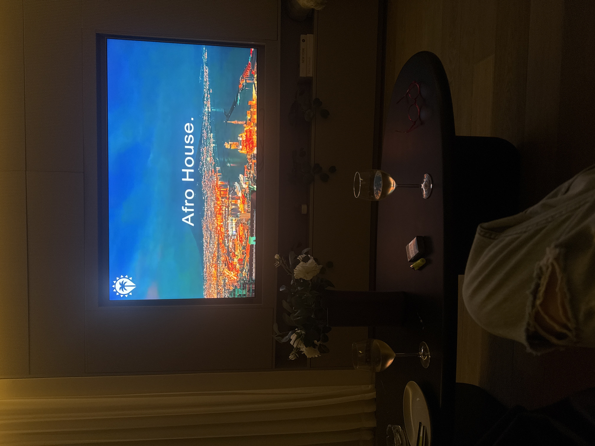 The image depicts an interior scene. A television screen displays the words "Afro House" over an image of a cityscape, possibly Tel-Aviv, Israel. Below the screen is a shelf or ledge holding a vase of white flowers. A dark-colored table sits in front of the screen with two wine glasses on it, one appearing full and the other partially filled. There are also eyeglasses and some small objects on the table. Portions of a person's legs and clothing are visible on the right edge of the image. In the bottom left corner, the edge of a curtain can be seen.