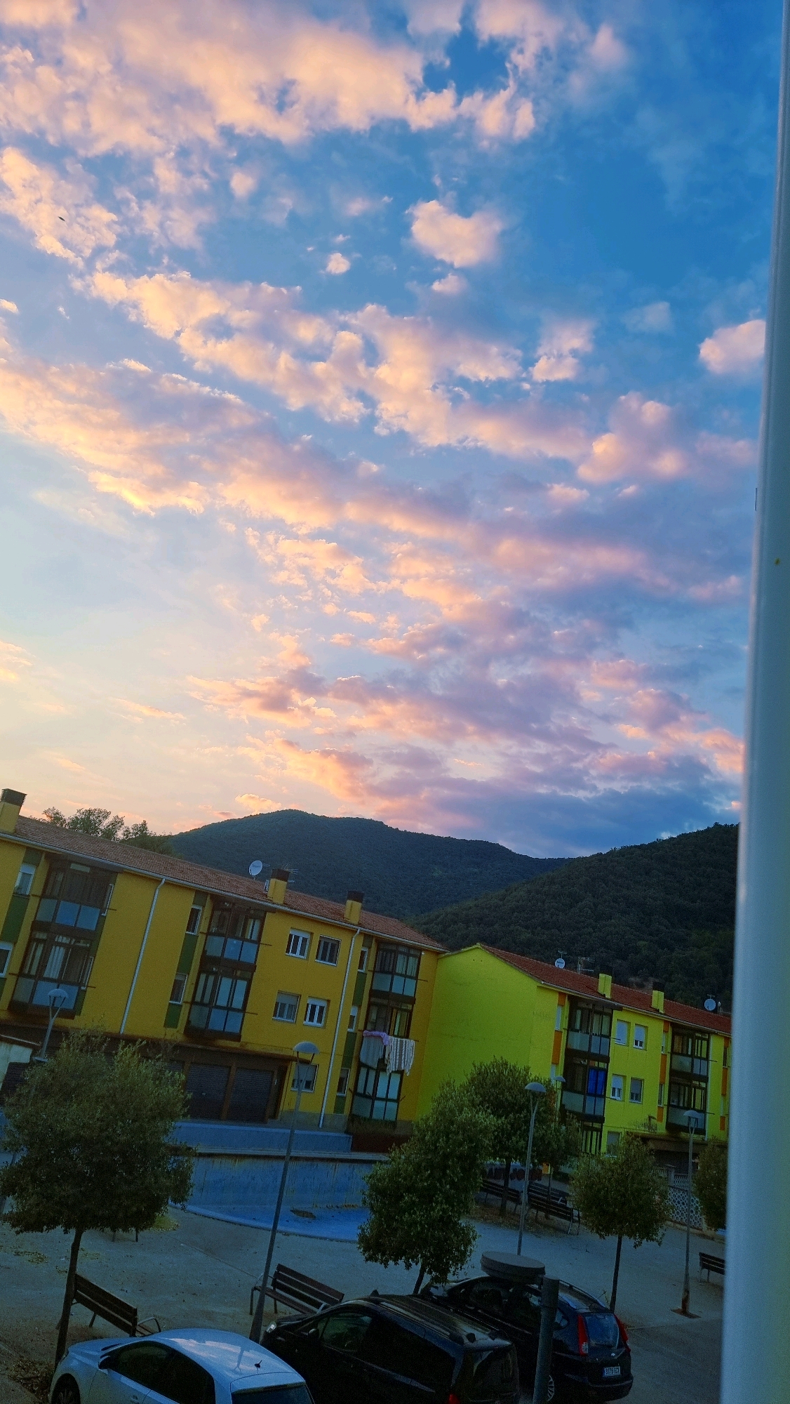 The image is an outdoor, eye-level shot of a residential area at dusk in Sant Joan Les Fonts, Spain. The sky is filled with a dramatic display of pink and orange clouds against a deep blue background, characteristic of a sunset or sunrise. In the foreground, a row of vibrant yellow apartment buildings with red-tiled roofs dominates the lower half of the frame. These buildings feature numerous windows and balconies, some with visible laundry hanging out to dry. A small courtyard area in front of the buildings contains trees, benches, and lampposts. Two cars are parked in the courtyard, one silver and one black. In the background, a dense, dark green forested mountain range rises under the colorful sky. The overall scene is serene, capturing the beauty of nature combined with urban living.